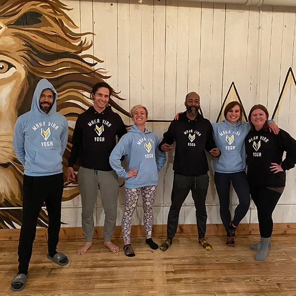 Group of six people standing in a yoga studio with a lion illustration on the wall behind them, all wearing matching Hoodies with 'Maha Vira Yoga' logo, smiling for the photo.
