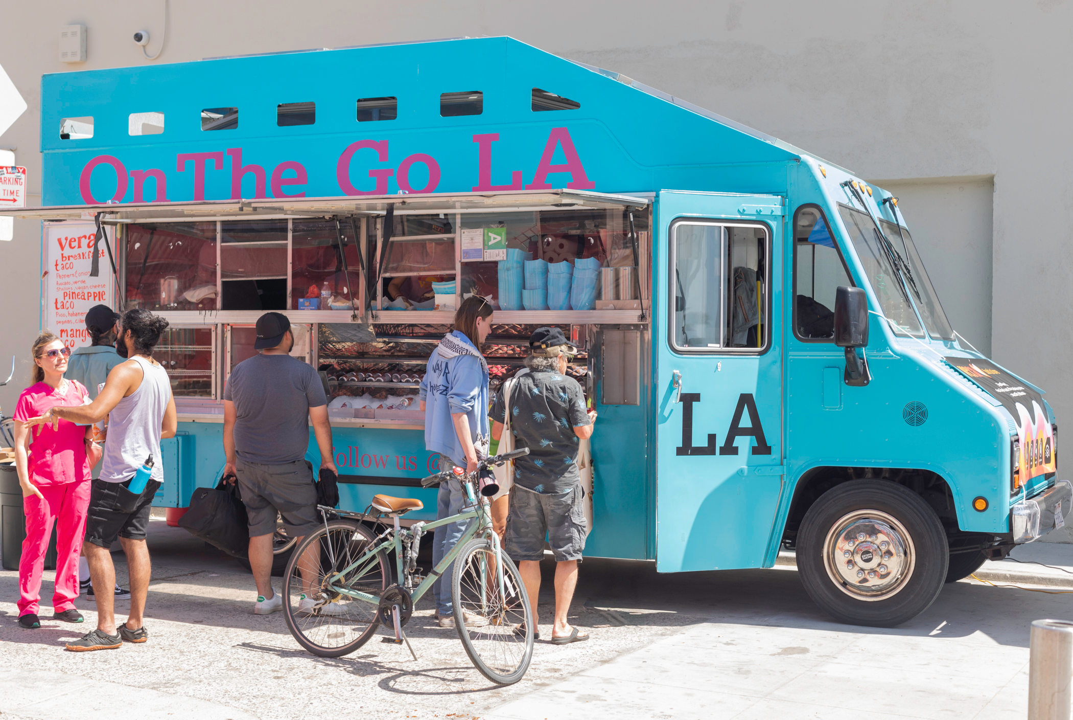 Blue food truck with pink signage has people gathered around and a person with a bicycle in front.
