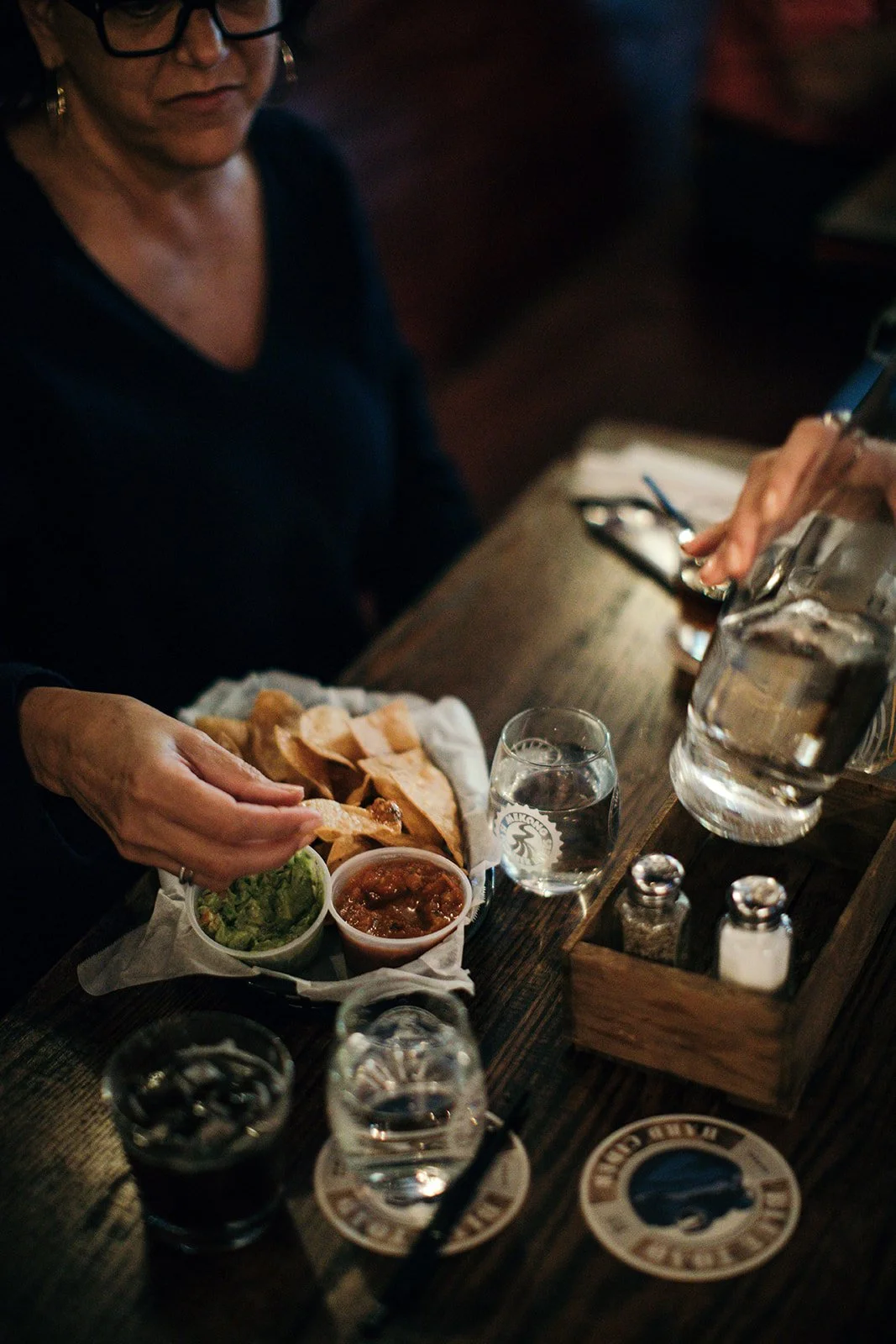 A woman sitting at a wooden table with a basket of tortilla chips, small containers of guacamole and salsa, a glass of water, and a pitcher of water. She is reaching for chips with her left hand, wearing glasses and a dark top.