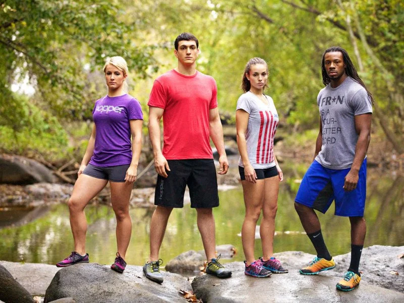 Four young adults standing on rocks by a creek in a wooded area, wearing activewear for outdoor activity.