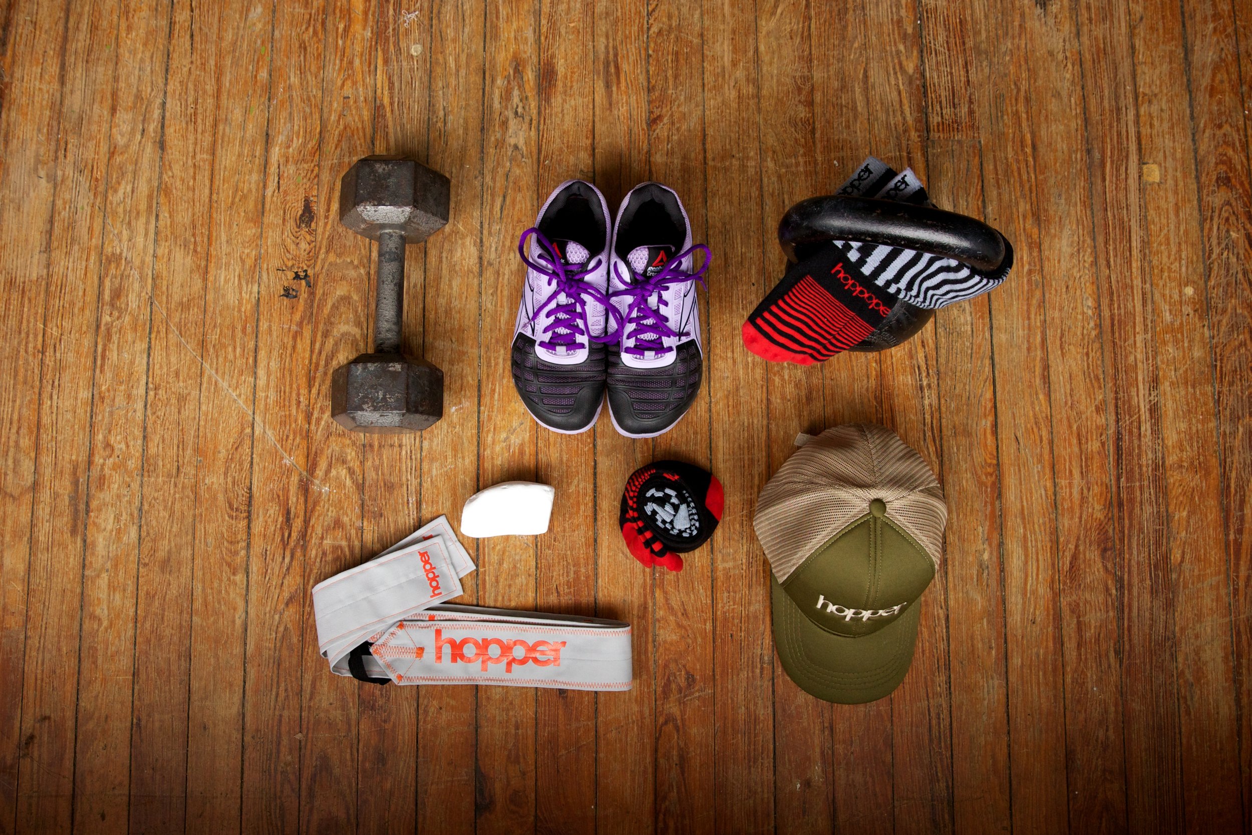 A wooden floor with a pair of purple and black athletic shoes, a black kettlebell, a small picnic-style hat, a black and striped cloth, a white towel with orange 'holler' branding, and a white and orange cloth bag with similar branding.