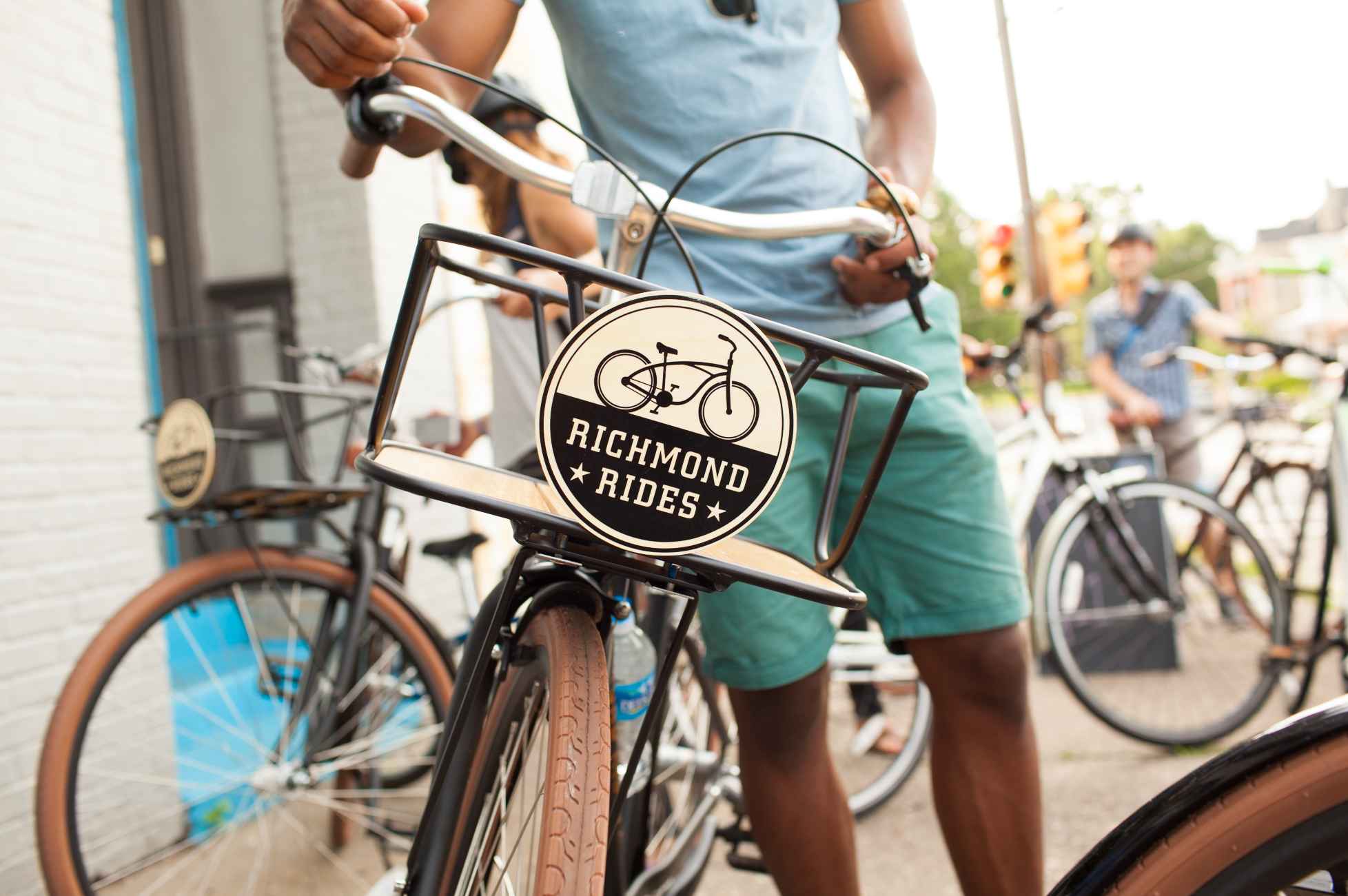 Close-up of a black bicycle with brown tires and a front rack that has a sign reading "Richmond Rides" with a bicycle illustration. There are people in the background, including a person in a blue shirt and green shorts holding the bike, standing out