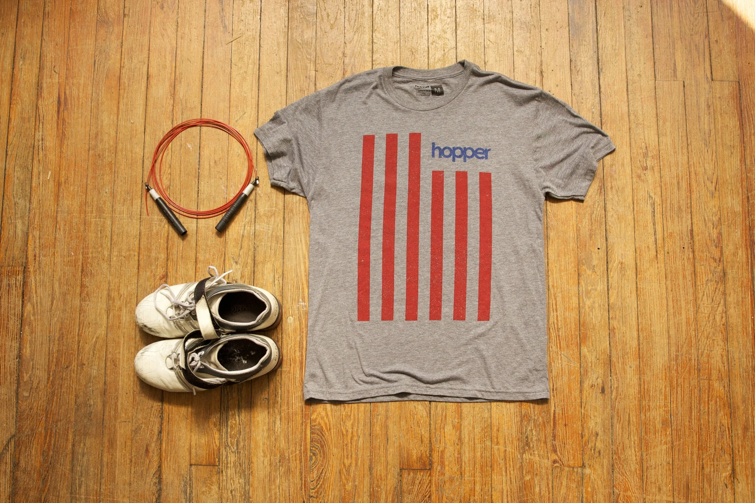 A gray T-shirt with red vertical stripes and blue text that says 'hopper,' placed on a wooden floor. To the left of the shirt, there is a jump rope with red handles, and at the bottom left, there is a pair of white athletic shoes.