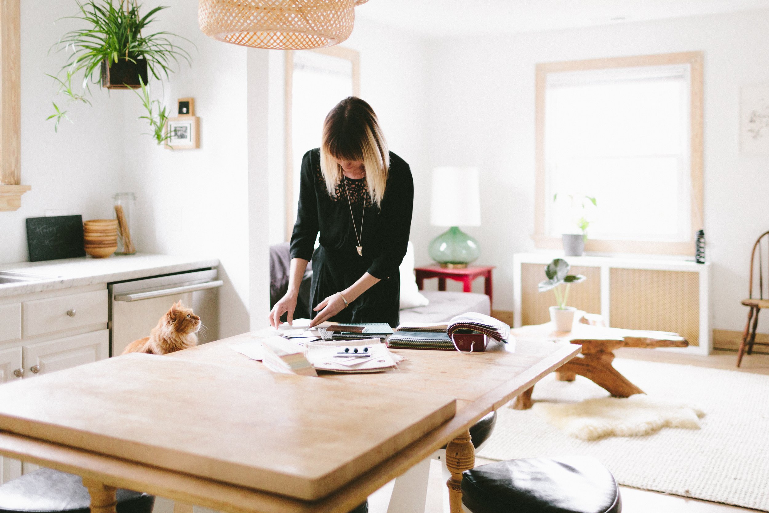 A woman with blonde-tipped hair working at a large wooden dining table with an orange tabby cat sitting nearby, in a bright, modern kitchen or dining area with natural light from windows, white walls, and various decor including plants, framed pictur
