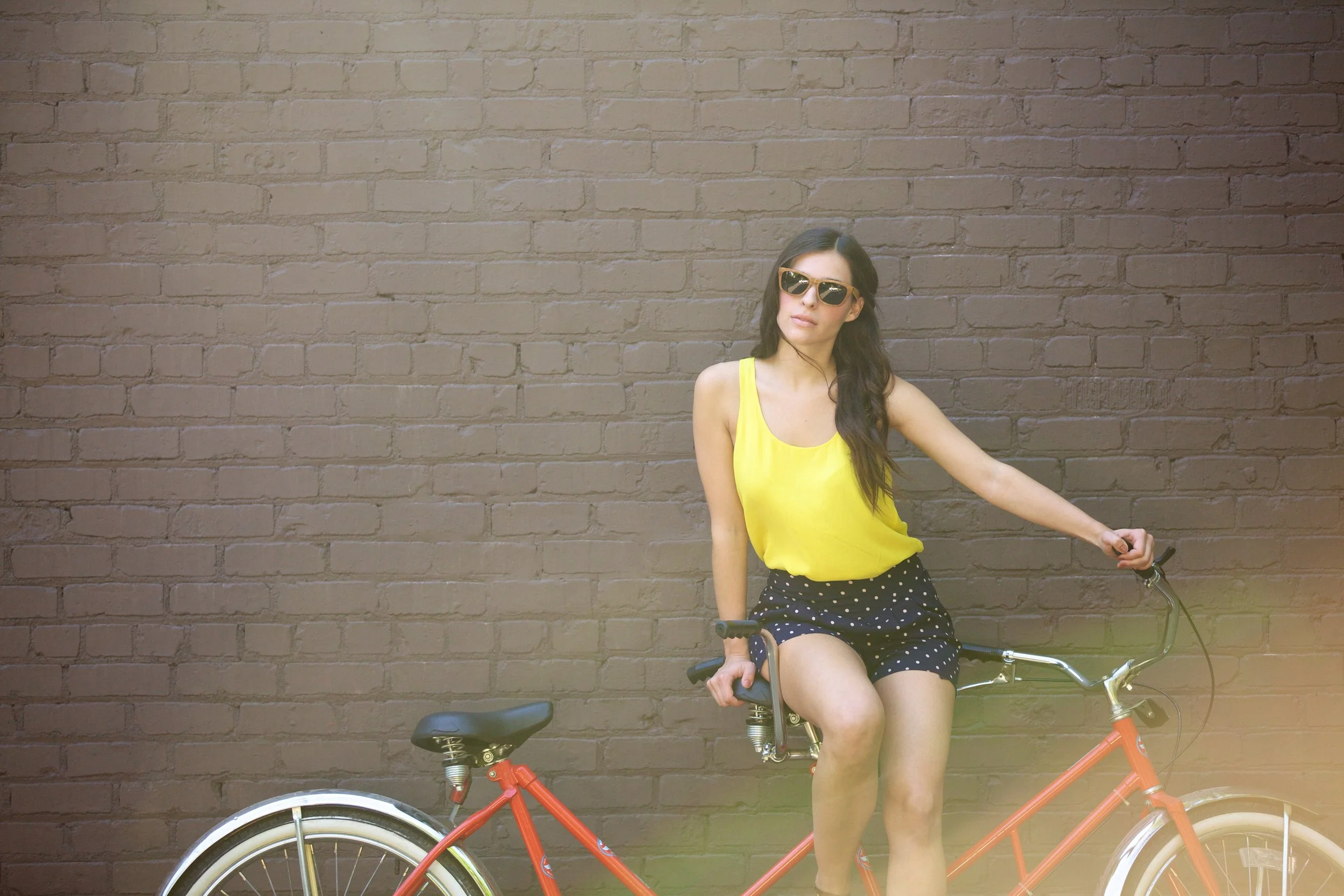 A woman wearing sunglasses and a yellow tank top sitting on a red bicycle against a brick wall.