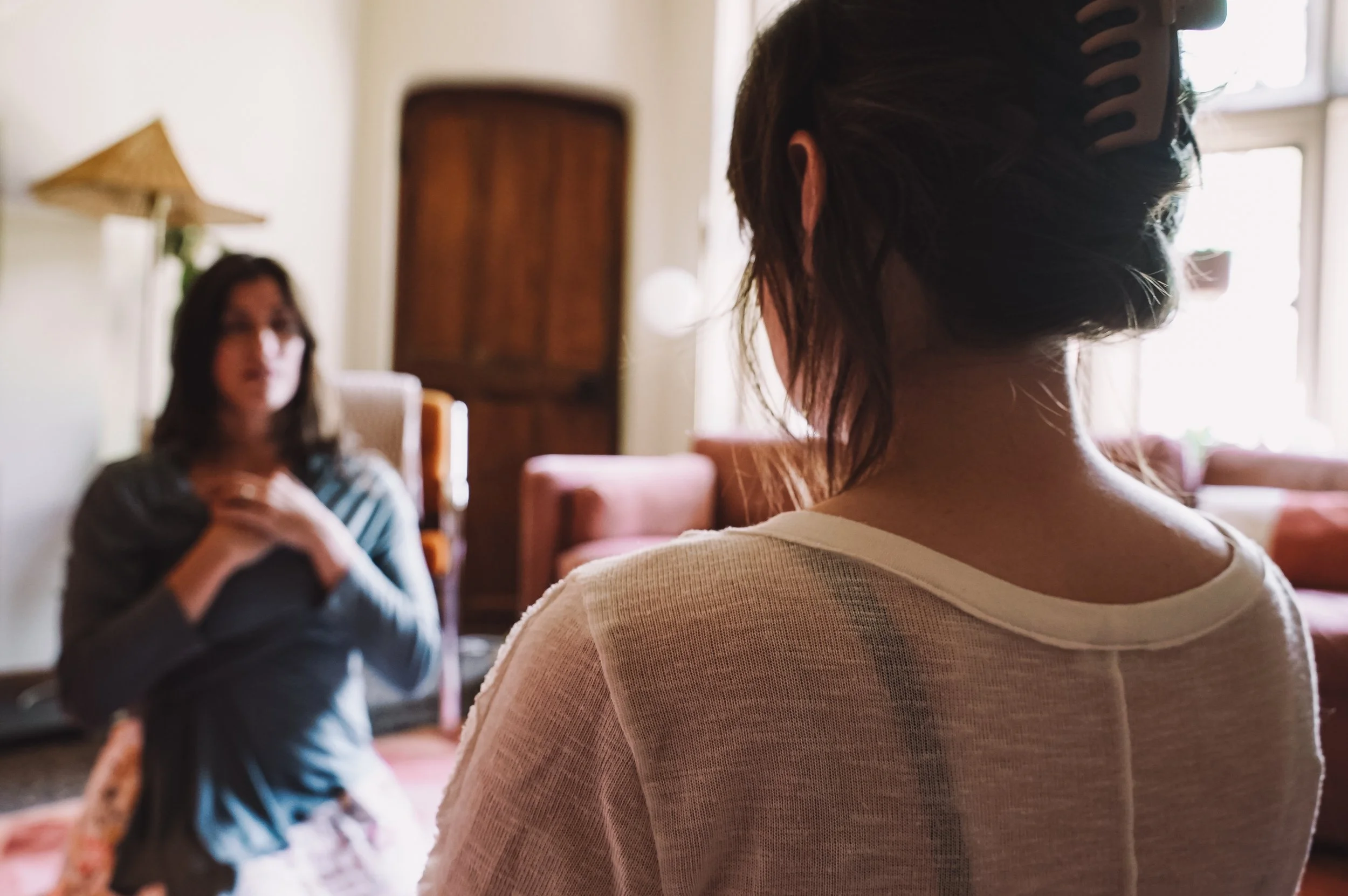 Two women engaging in a conversation indoors with natural light from windows. The woman in the foreground has dark hair tied back with a hair clip, wearing a light-colored shirt. The woman in the background has shoulder-length hair, wearing a striped