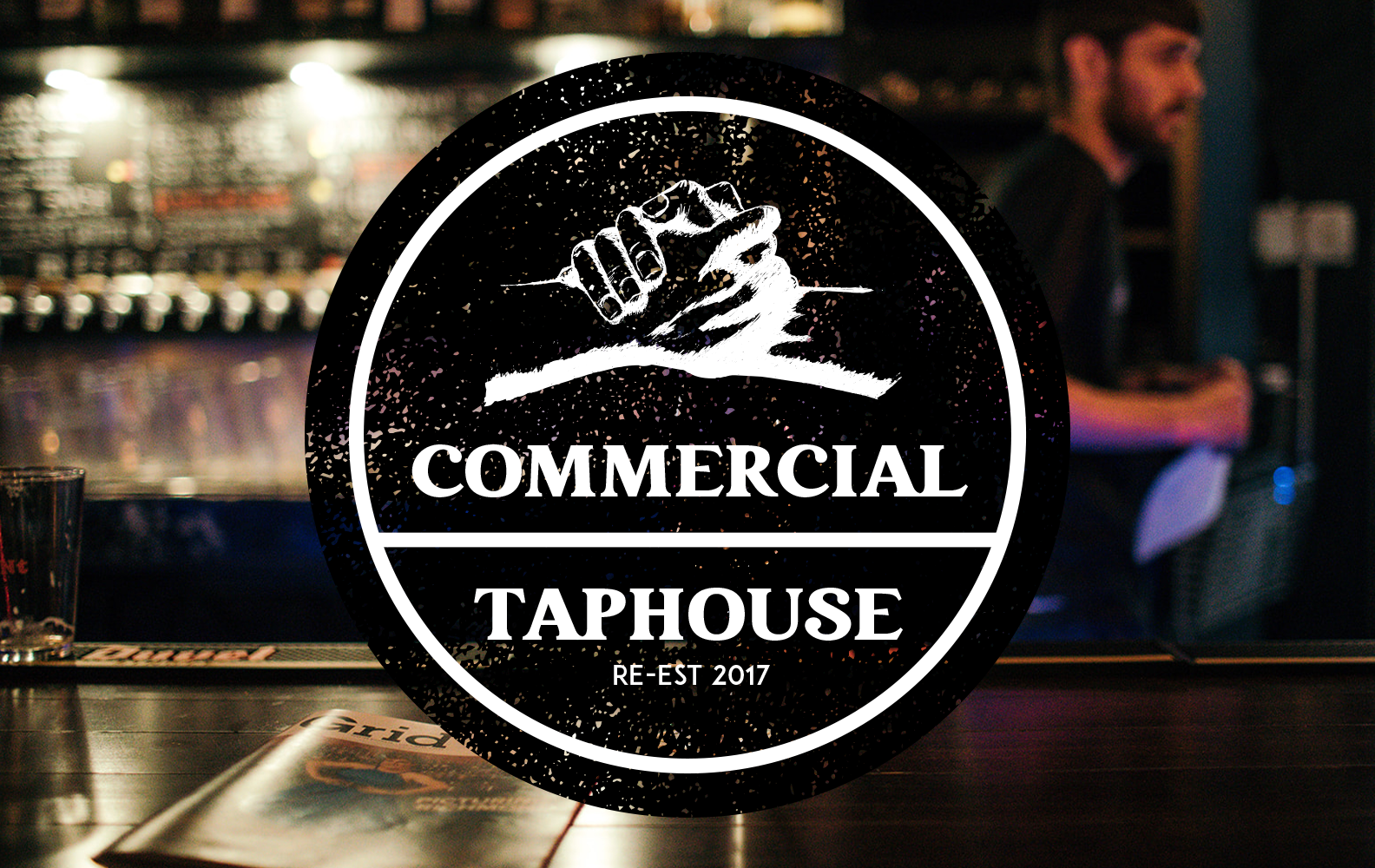 Logo of a commercial taphouse featuring a drawn fist bump, with text 'Commercial Taphouse, Re-Est 2017' over the background of a bar with a bartender.