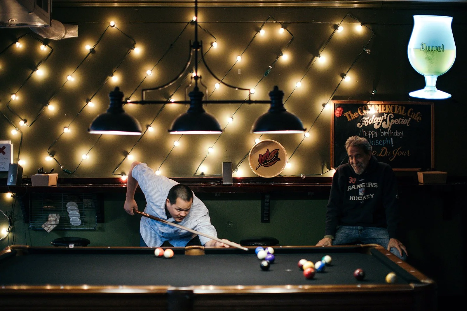 Two men playing pool in a dimly lit bar with string lights on the wall and a blackboard with birthday wishes behind them.