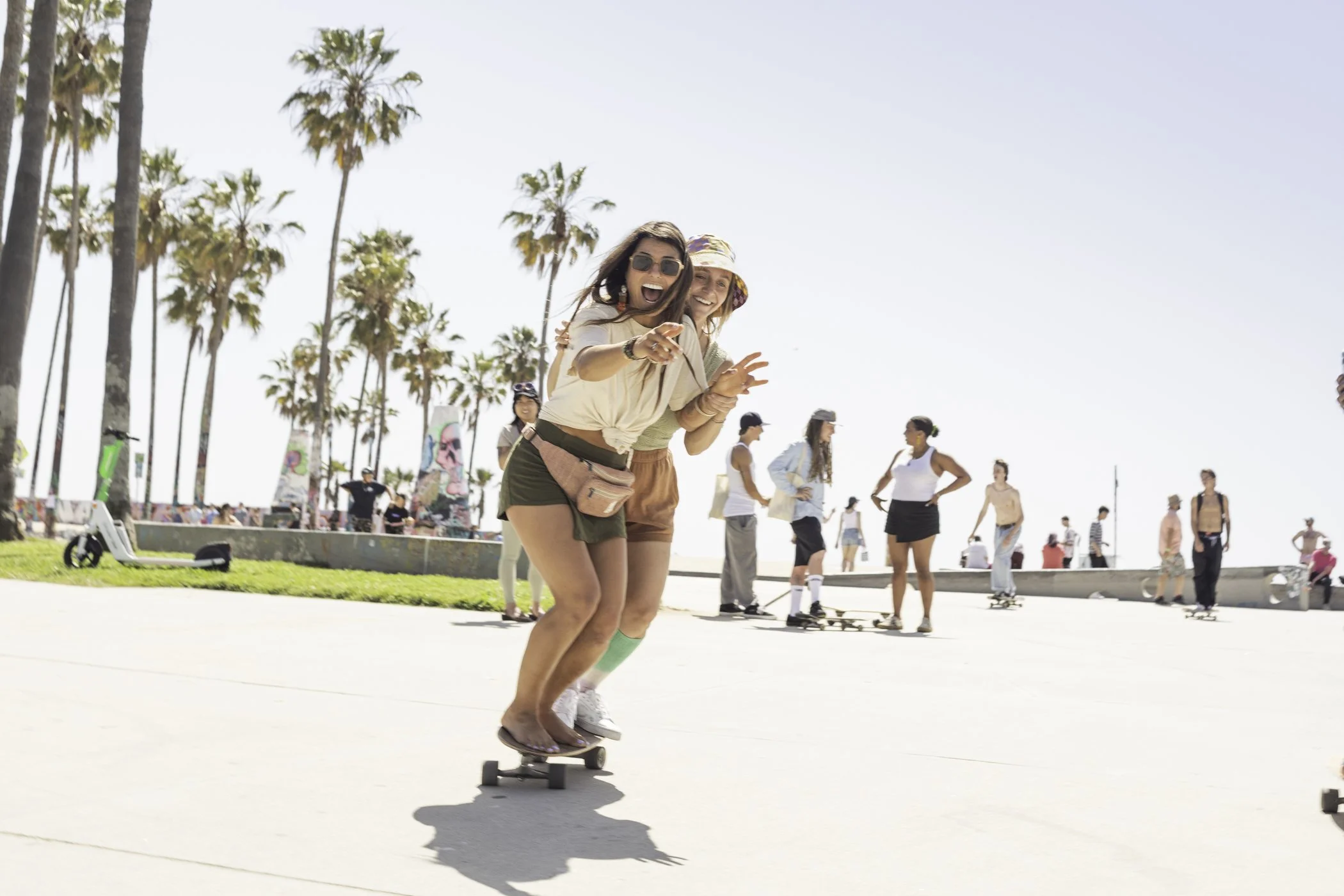 Two young women skateboarding on a sunny beachside promenade, with palm trees and people in the background enjoying the day.