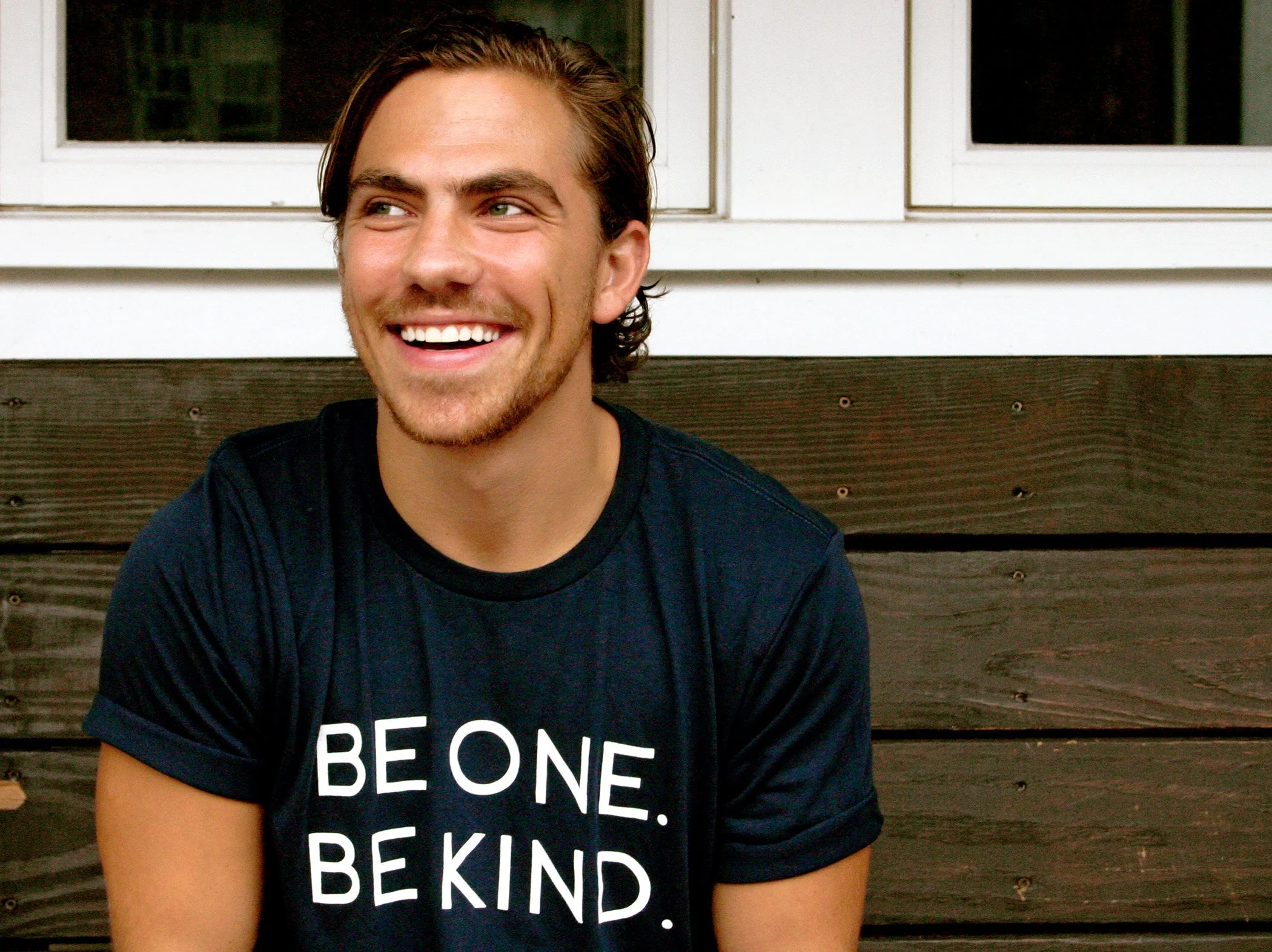 A young man with brown hair and a beard sitting in front of a house with white window frames and a brown wooden fence, smiling and wearing a black t-shirt that says 'BE ONE. BE KIND.'