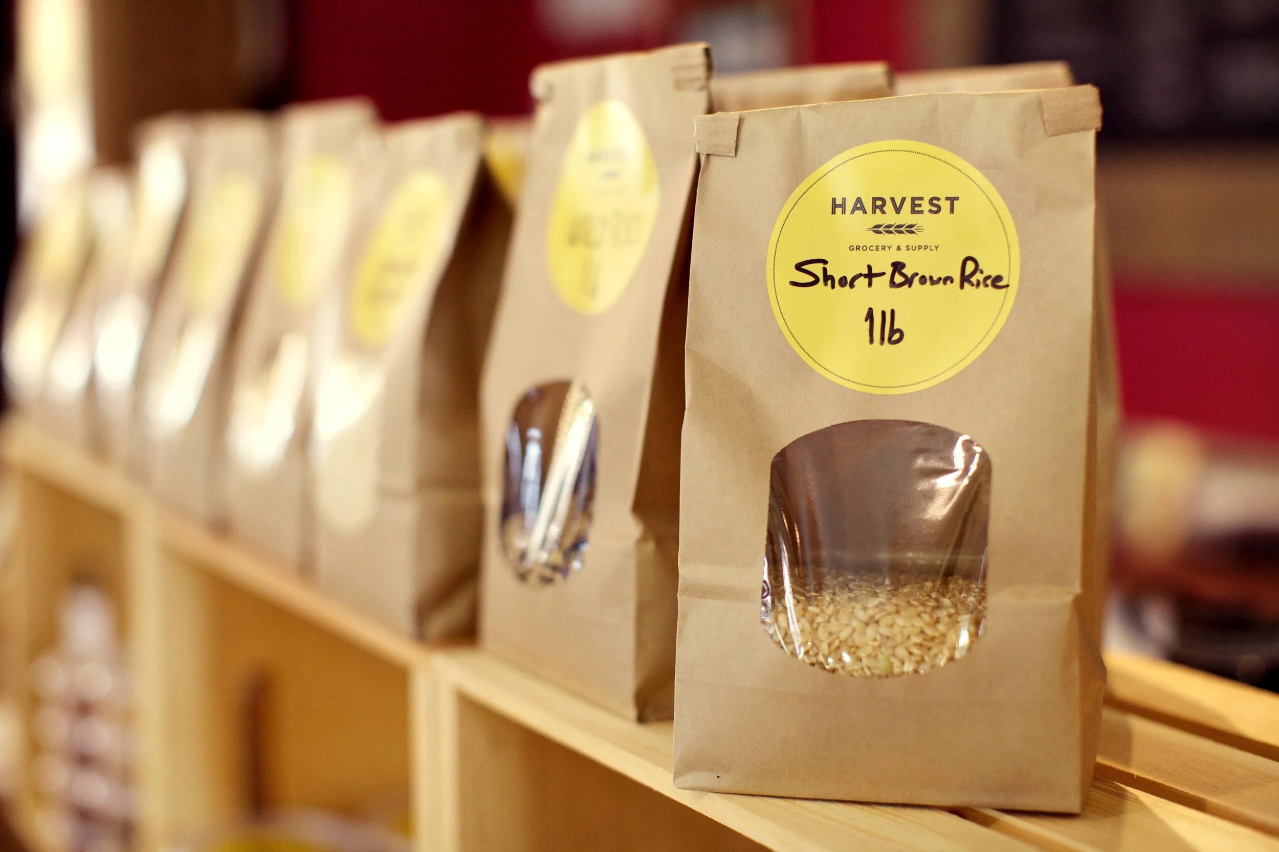 Bundles of short brown rice packaged in brown paper bags with yellow labels on a wooden shelf.