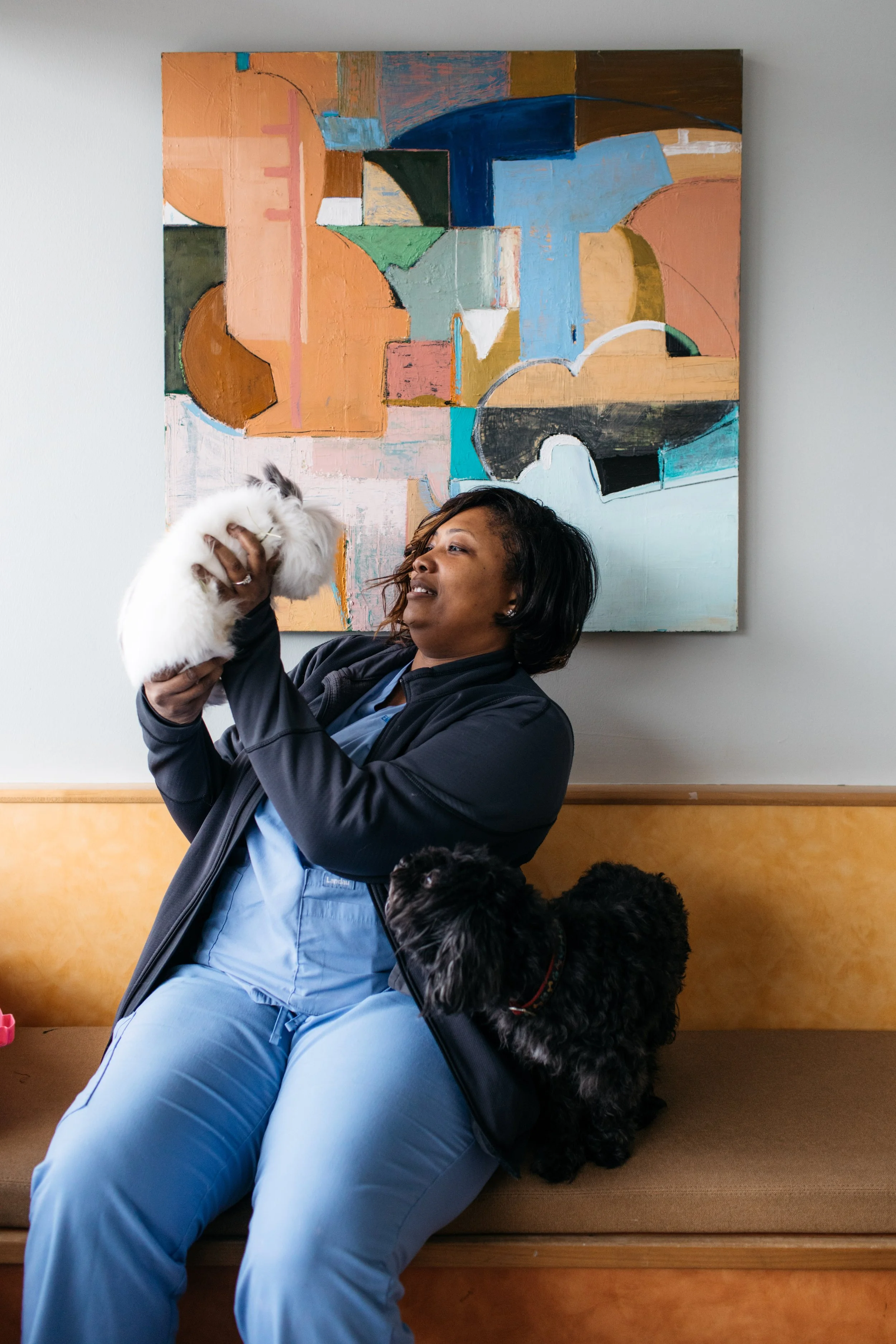 A woman holding a white rabbit in her hands, sitting on a wooden bench in front of a colorful abstract painting, with a black dog sitting beside her.