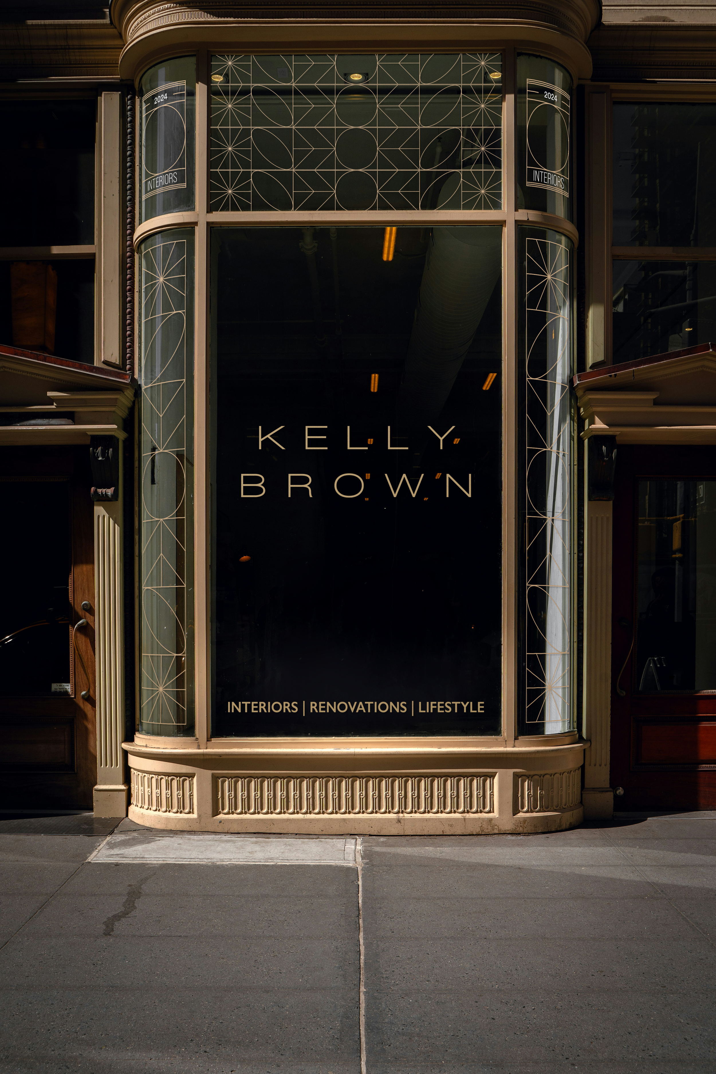 Front storefront of Kelly Brown, a business specializing in interiors, renovations, and lifestyle. The storefront features a large window with gold lettering and decorative trim, and the sidewalk in front is concrete with visible cracks.