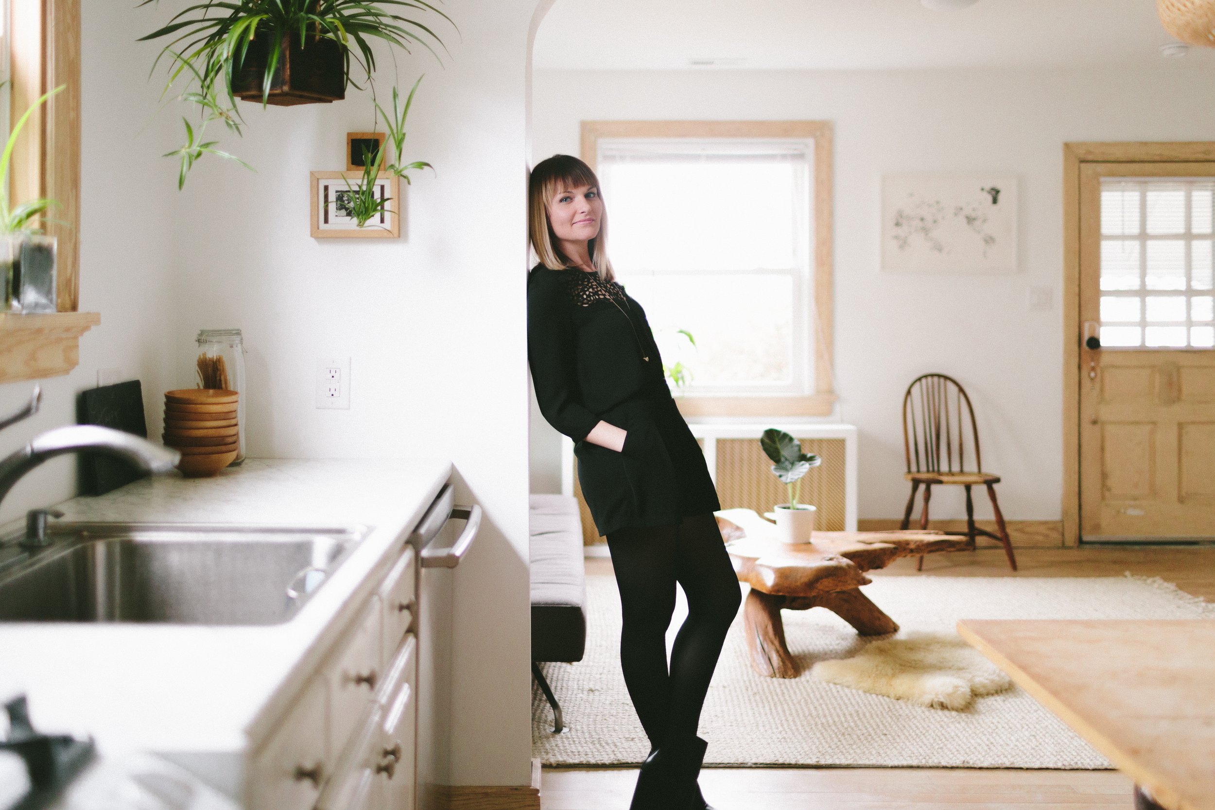 A woman with shoulder-length hair and bangs, smiling, wearing a black outfit, standing in a bright, cozy room with wooden accents, plants, and a door with glass panes.