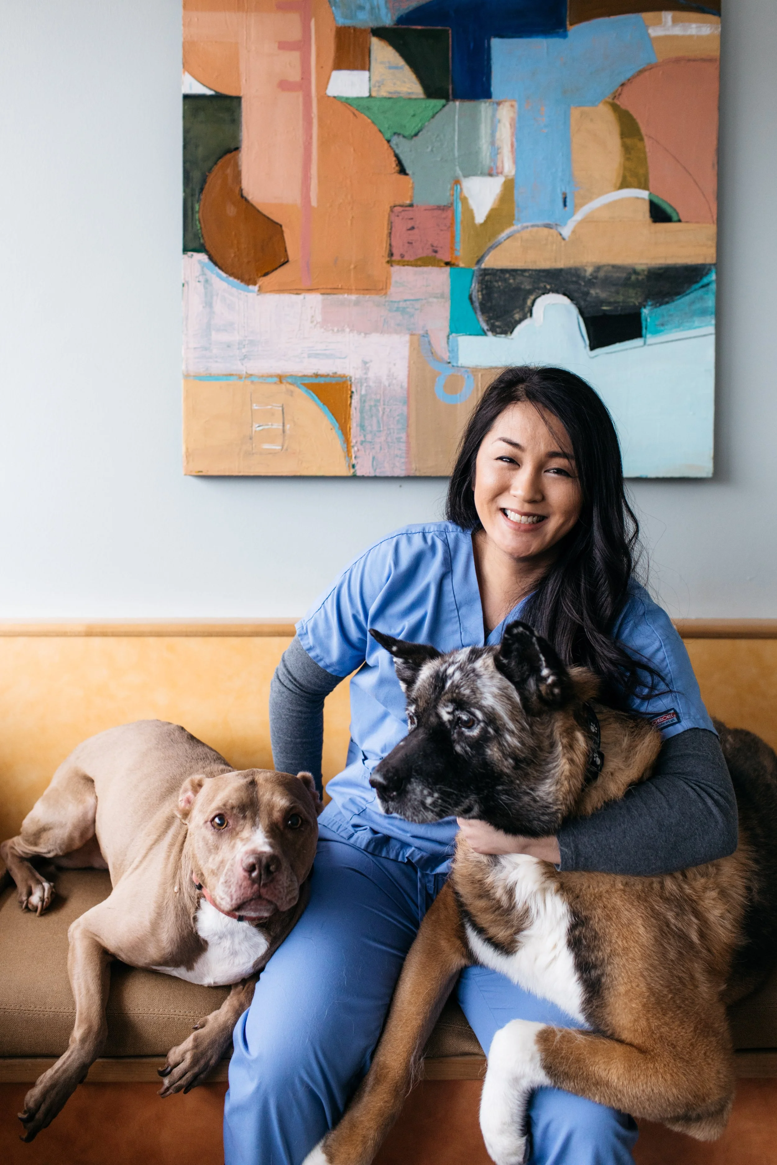 A woman in blue scrubs smiling while holding two puppies—a merle Australian Shepherd and a German Shepherd—sitting on a beige couch with a wooden trim, with a colorful abstract painting hanging on the wall behind her.