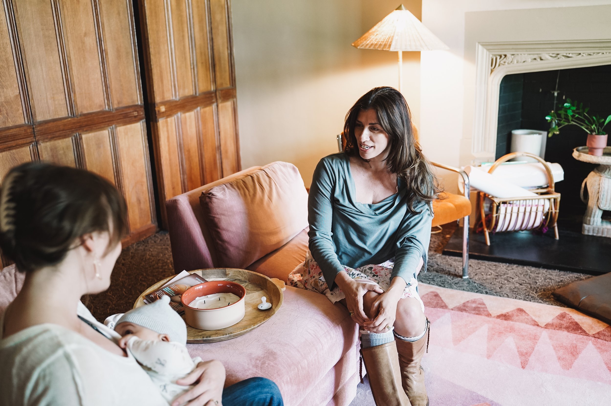 A woman with long dark hair sitting on a pink sofa, smiling while talking to another woman holding a baby. The scene is in a cozy living room with wood-paneled walls, a lit lamp, a fireplace with a plant on the mantle, and a wicker side table with a 