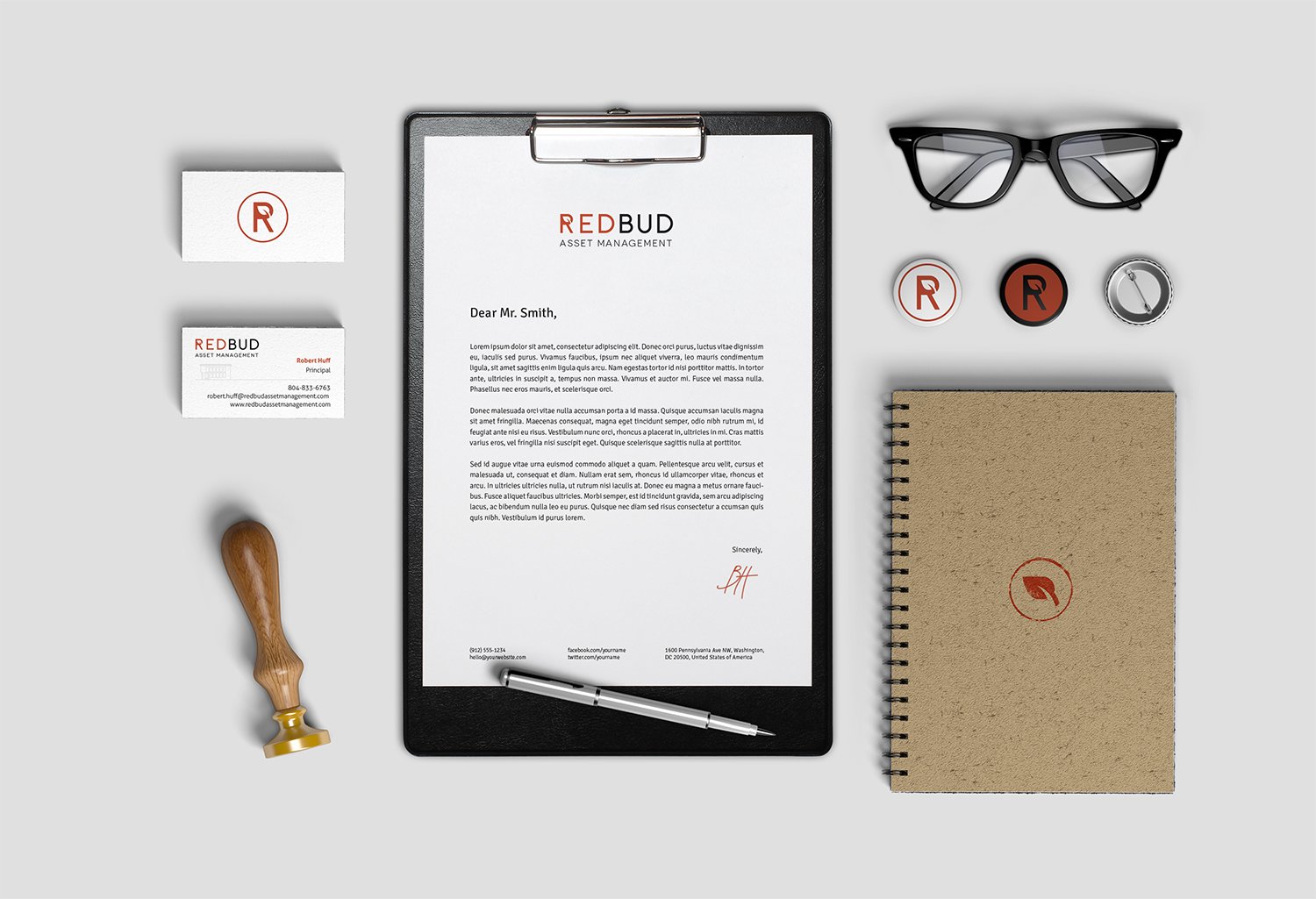 A collection of office supplies and branded materials including a letter on a clipboard, business cards, a rubber stamp, a stamp pad, a pair of black eyeglasses, a notebook with a circular logo, a pen, and some branded buttons, arranged on a light gr