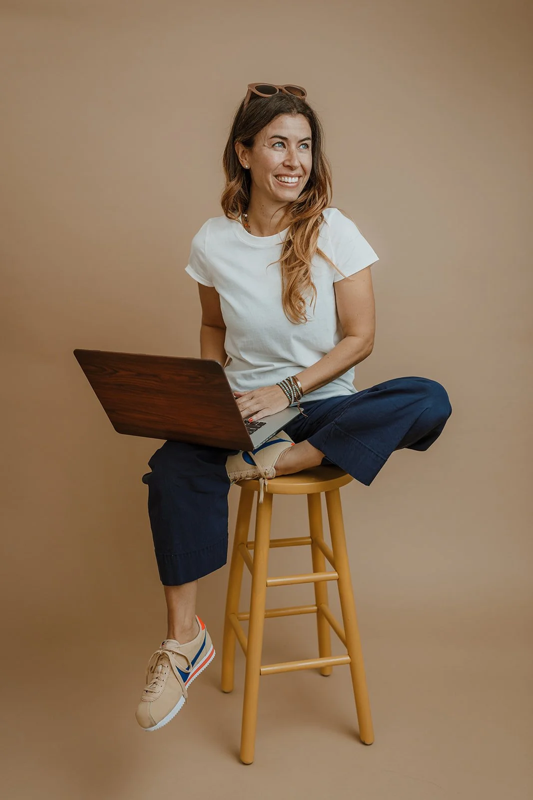 A woman with long brown hair, wearing a white t-shirt, dark blue pants, and beige sneakers, sits cross-legged on a wooden stool with a laptop on her lap, smiling and looking to her right against a plain beige background.