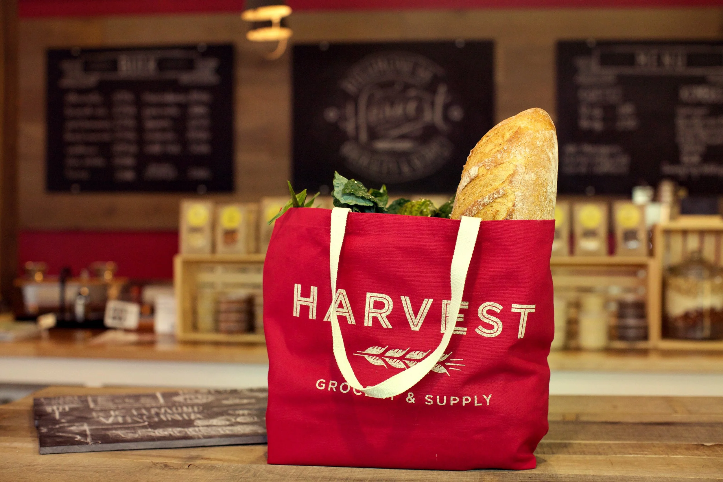 Red tote bag with white text and a leaf logo, filled with a baguette and greens, on a wooden table in a cafe or grocery store.