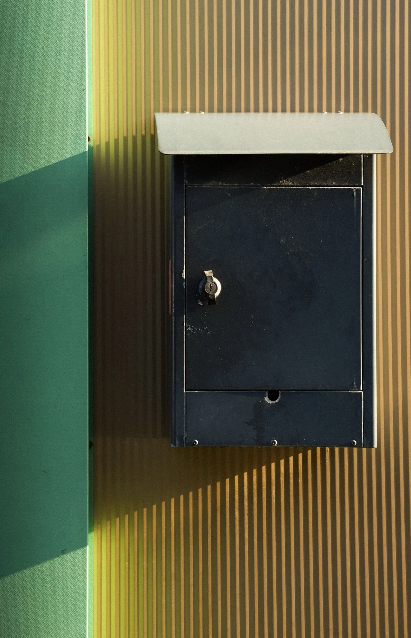 Black mailbox with a silver lid and a lock, mounted on a textured wall with green and yellow stripes.