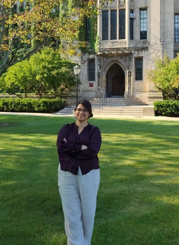 woman with dark hair, black sweater, and light jeans standing on grass in front of stone building. Arms crossed.