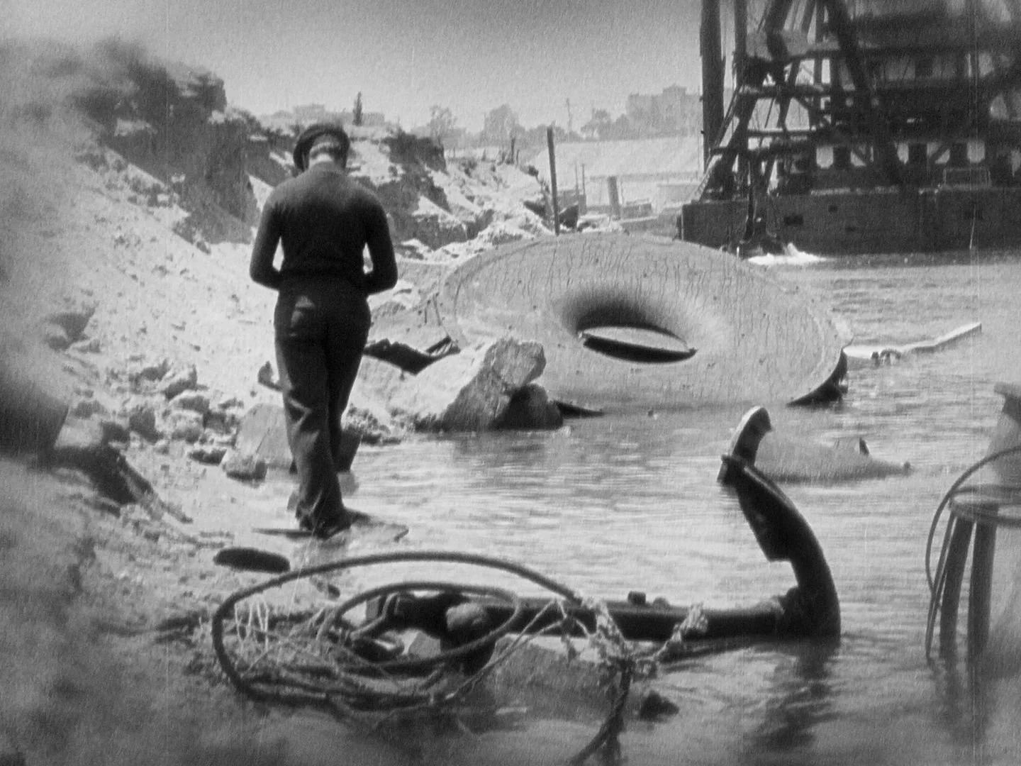 Woman walking along a shoreline with debris, including a large pipe and part of a broken ship, in a coastal area with industrial structures in the background.