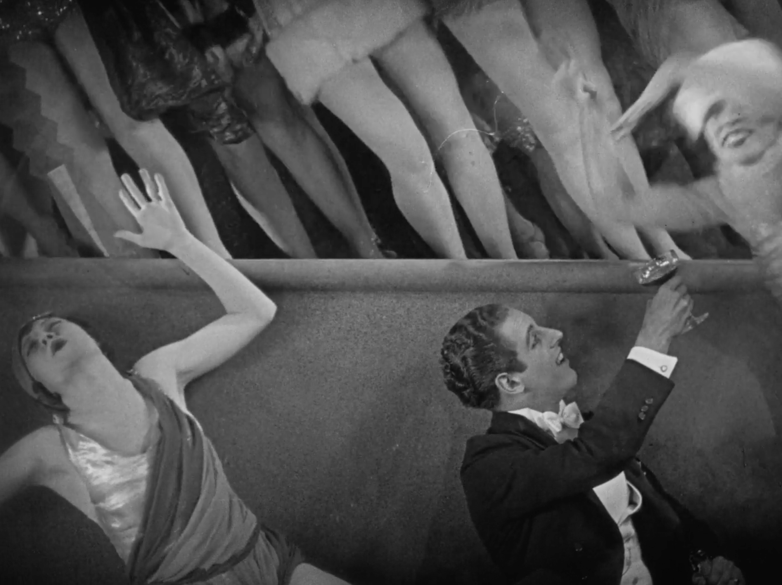 In a black and white photo, two women and one man are sitting at a table, holding drinks and engaging in conversation. The women have their heads tilted back, with one raising her hand. The men are dressed in tuxedos, and the scene appears to be lively and social.