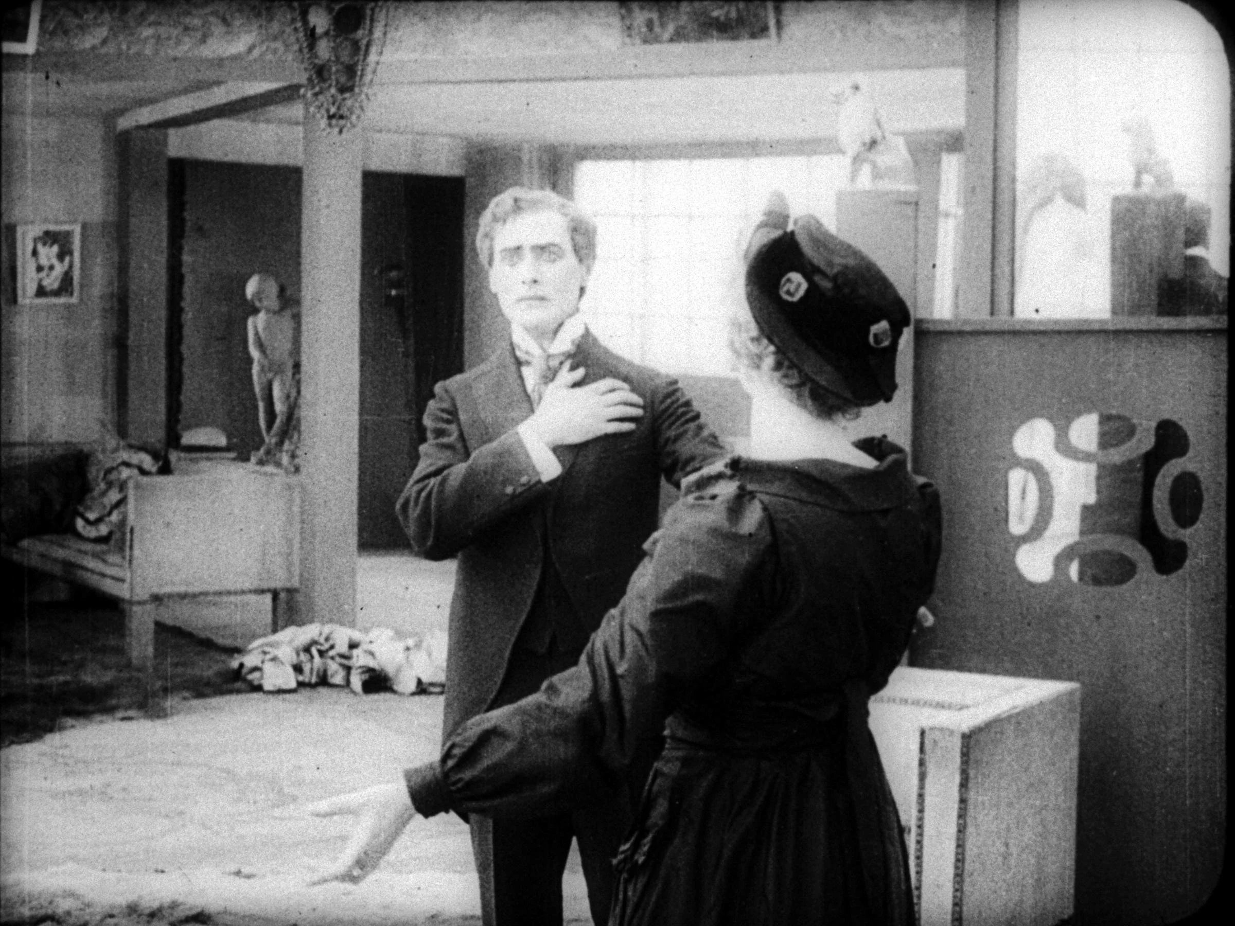 A black and white photo of a man in a suit with a woman in a dark dress and hat, in an art gallery or museum setting. The man appears to be touching his collar or chest while the woman gestures with her hand. Artwork and sculptures are visible in the background.