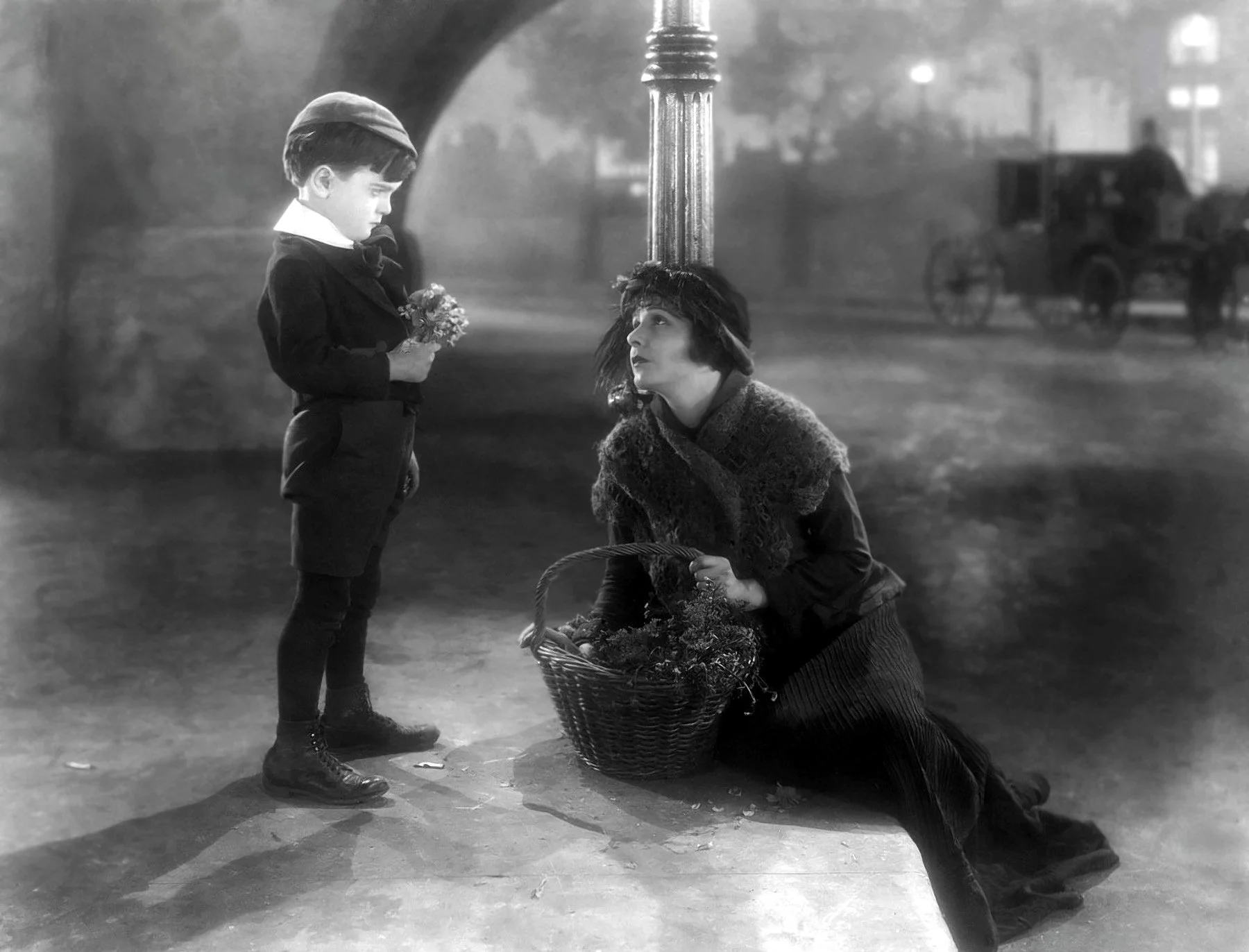 A young boy standing on a street, holding flowers, looks down at a kneeling woman holding a basket, who is gazing up at him. The scene appears nostalgic and emotional, with a blurred background of a street scene and a horse-drawn carriage.