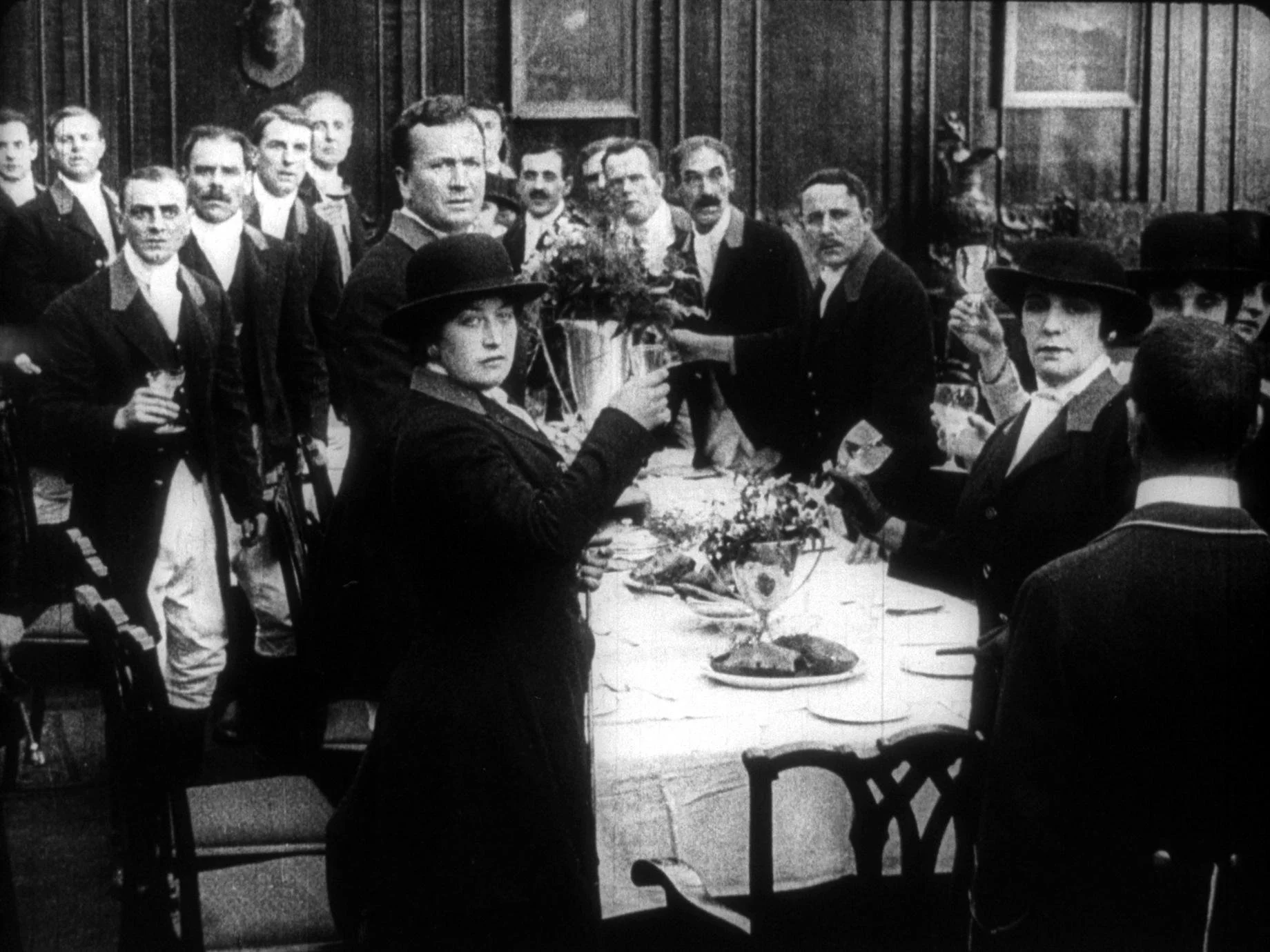 A black and white photograph of a group of people gathered around a table with food and flowers, with some individuals holding glasses. The scene appears to be a formal or celebration event in a vintage setting.