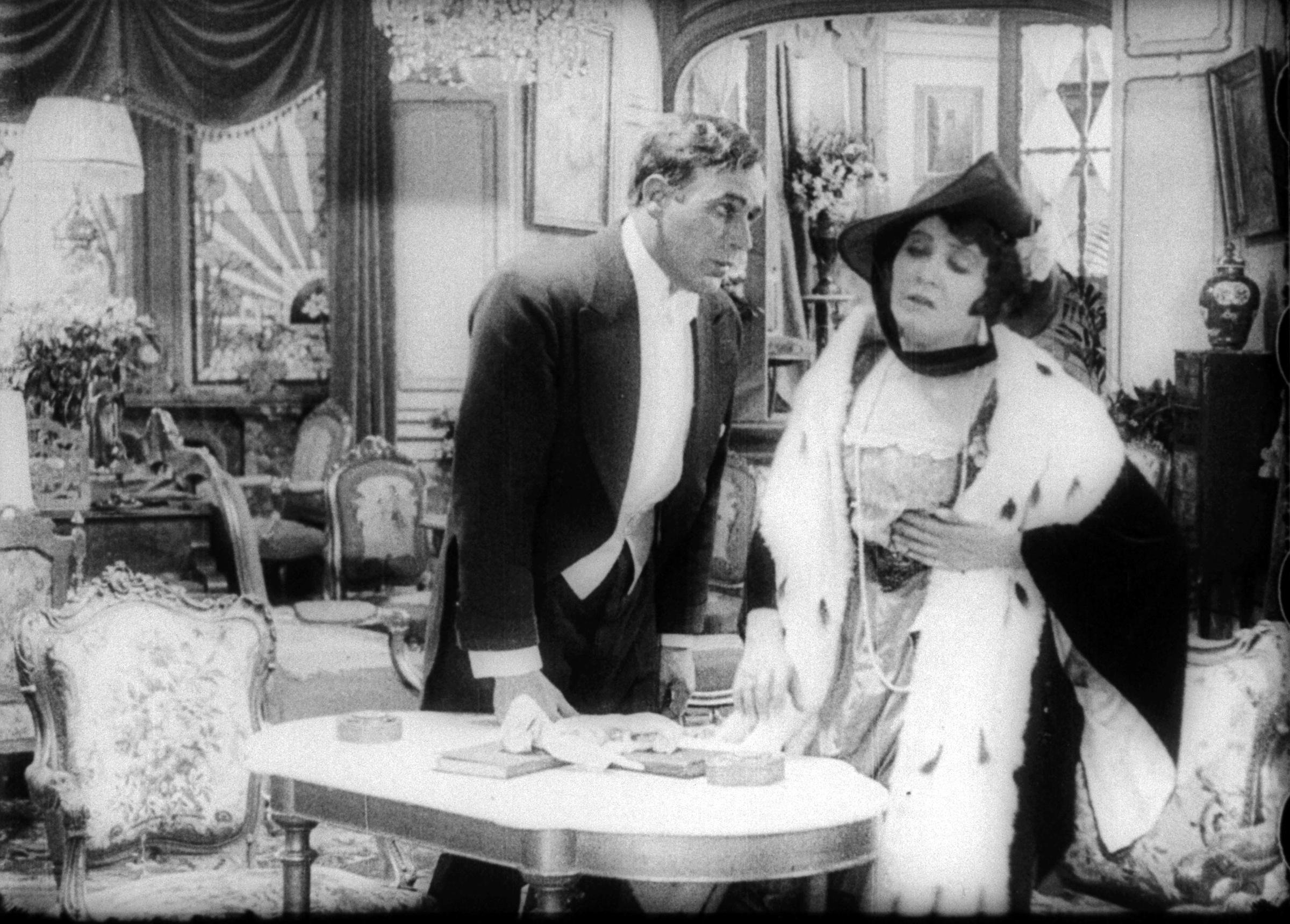 A black-and-white scene from a theatrical or film performance showing a man in formal wear and a woman in a costume with a hat, in an elegant room with ornate furniture and decor.
