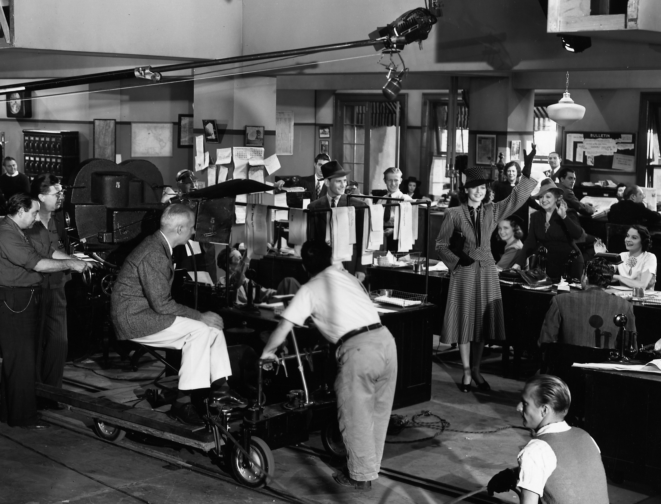 Black and white photograph of a film or television studio set, resembling a 1930s office, with people dressed in period clothing. A woman in a striped suit and hat is waving to the camera, surrounded by various crew members and actors, with filming equipment and lighting visible.