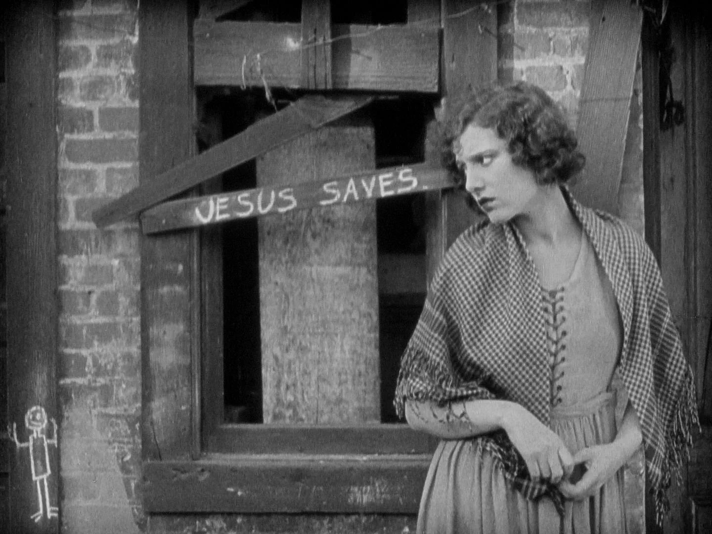 A woman with curly hair looking to the side, standing in front of a rustic wooden structure with a sign that reads 'JESUS SAVES' in a black-and-white setting.