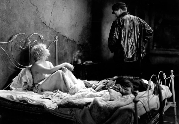 A woman with curly hair lying in bed, looking at a man standing beside her bed in a dark room with a textured wall.