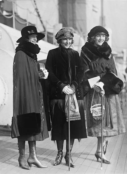Three women dressed in vintage winter clothing, standing outdoors with a ship in the background.