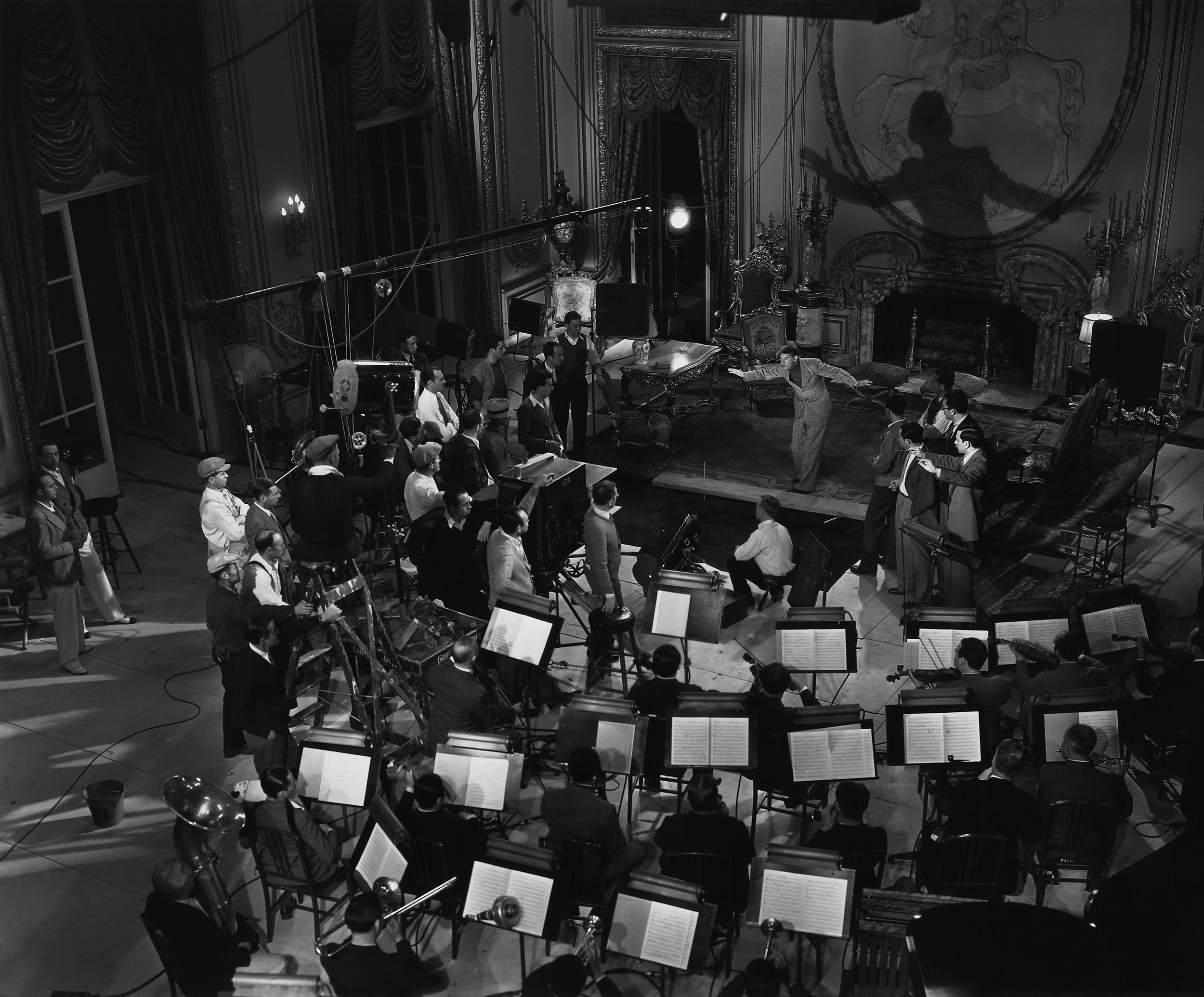 Black and white photograph of a theater stage with a performer and a group of people, including an orchestra, crew, and audience members, preparing for a performance.