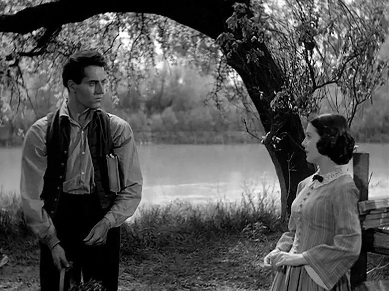 A black-and-white still from a movie showing a young man and woman standing outdoors near a river, under a large tree. The young man holds books and a textbook, and the woman is dressed in vintage clothing with a bow tie, standing near a bench with stacked books.