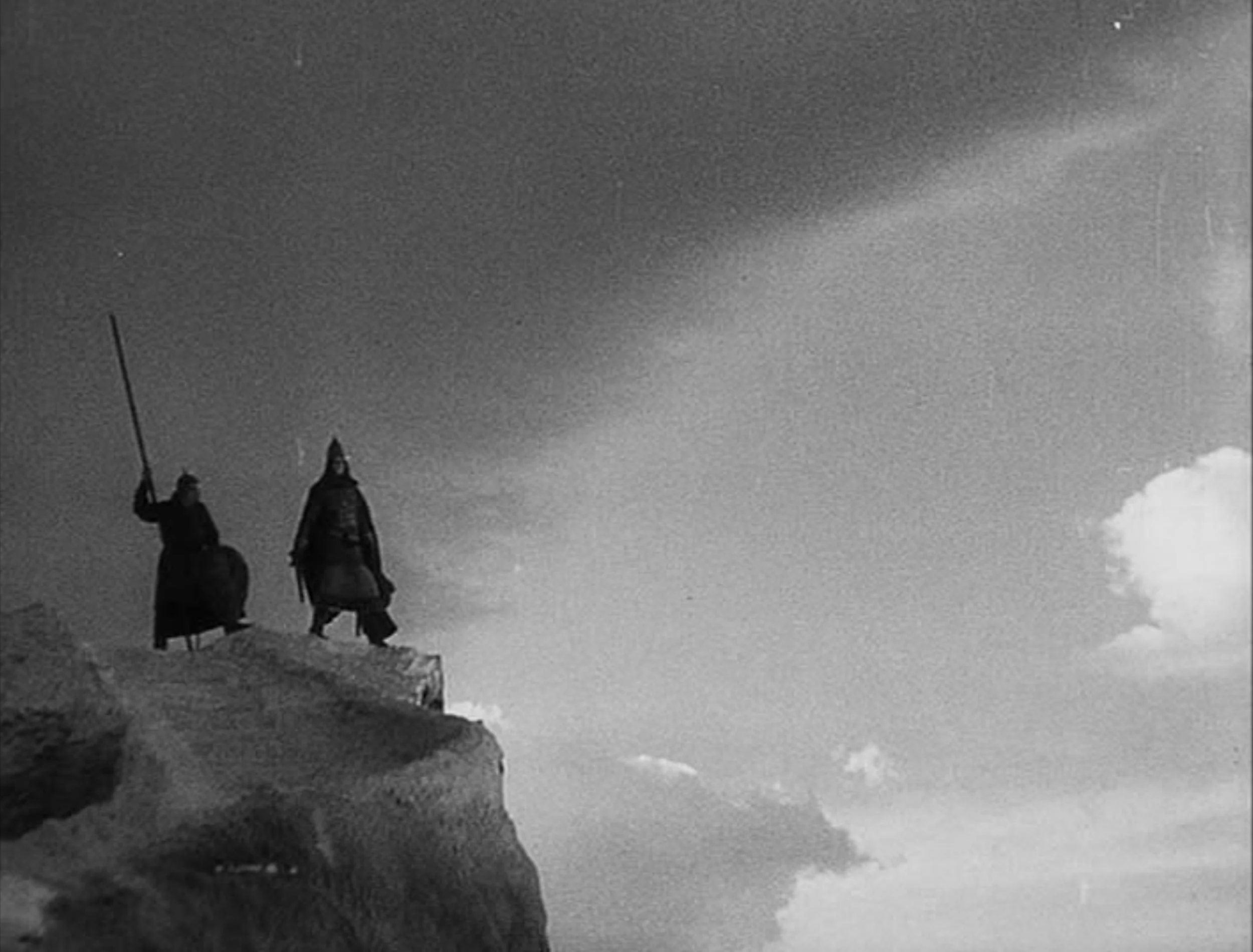 Two monks standing on a rocky mountaintop, dressed in dark robes, with one holding a staff against a cloudy sky.