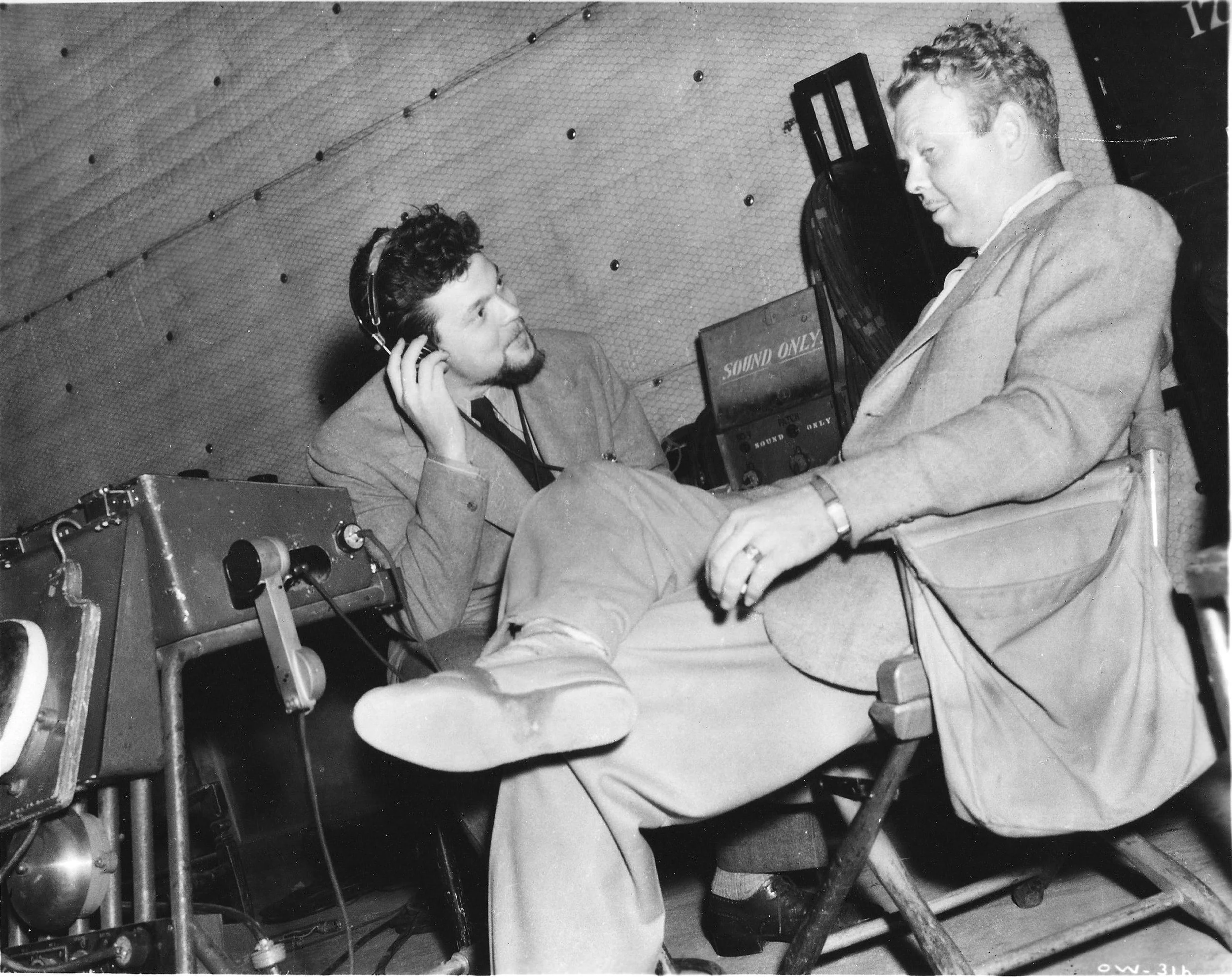 Black and white photo of two men in conversation. One man is sitting on a chair with legs crossed, wearing a suit and show, and is listening with headphones. The other man is lying on a table or bed with his legs propped up, dressed in a suit, and gesturing with his hand.