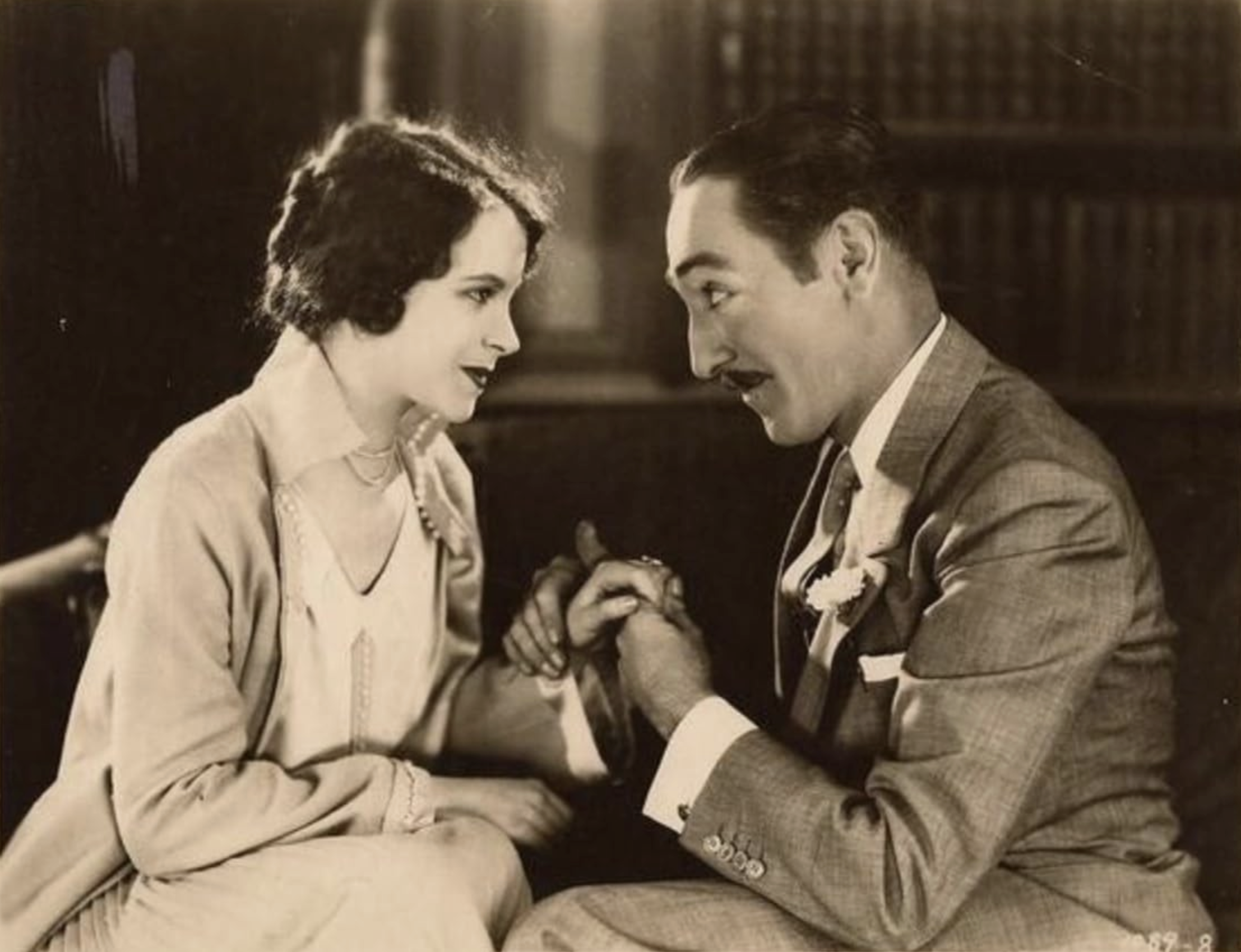 A black and white photo of a woman and a man sitting close together, holding hands and smiling at each other in a room with bookshelves in the background.