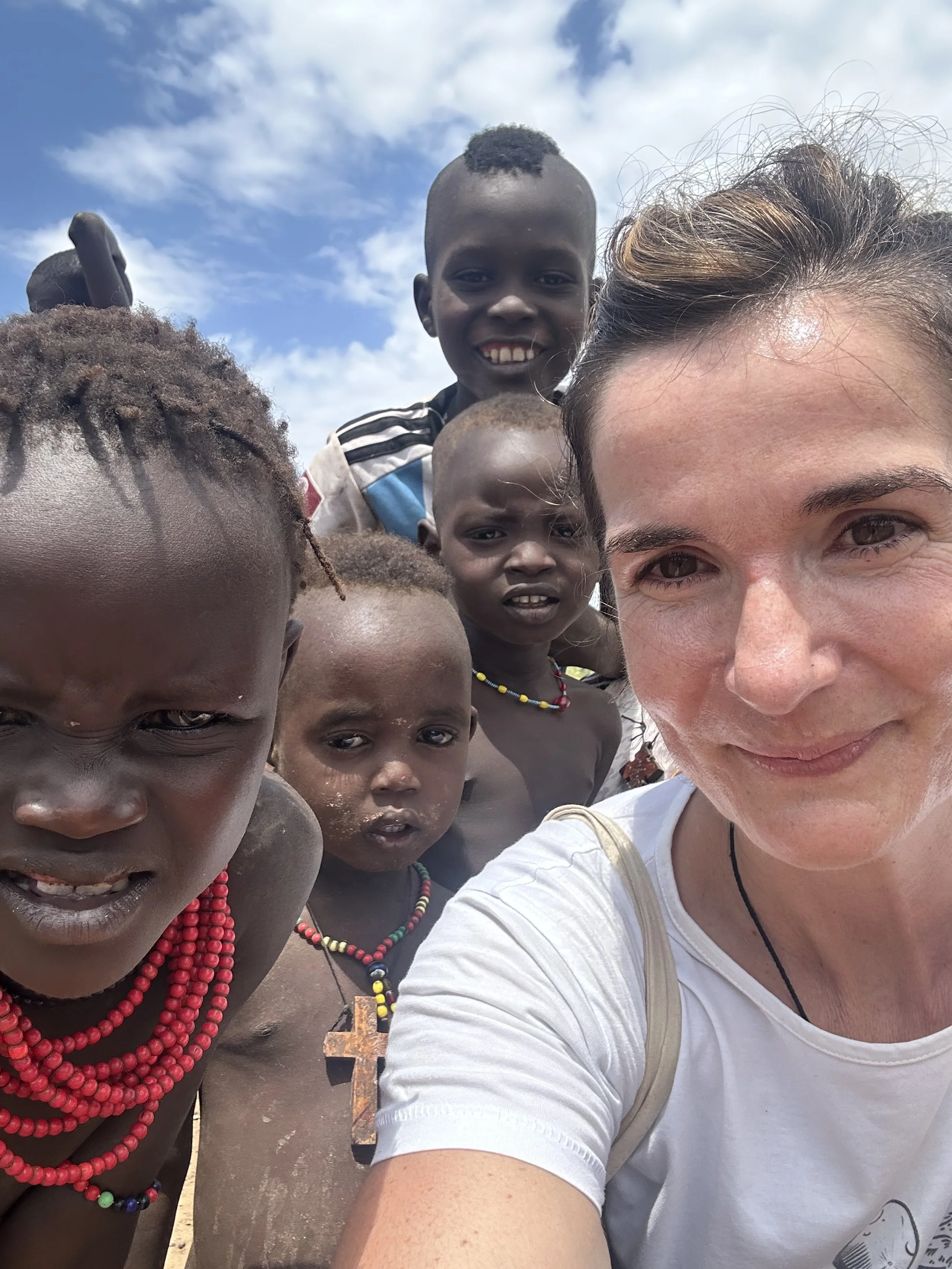 A woman taking a selfie with six children in a rural outdoor setting under a blue sky with some clouds. The children are wearing colorful beaded necklaces and appear to be of African descent.