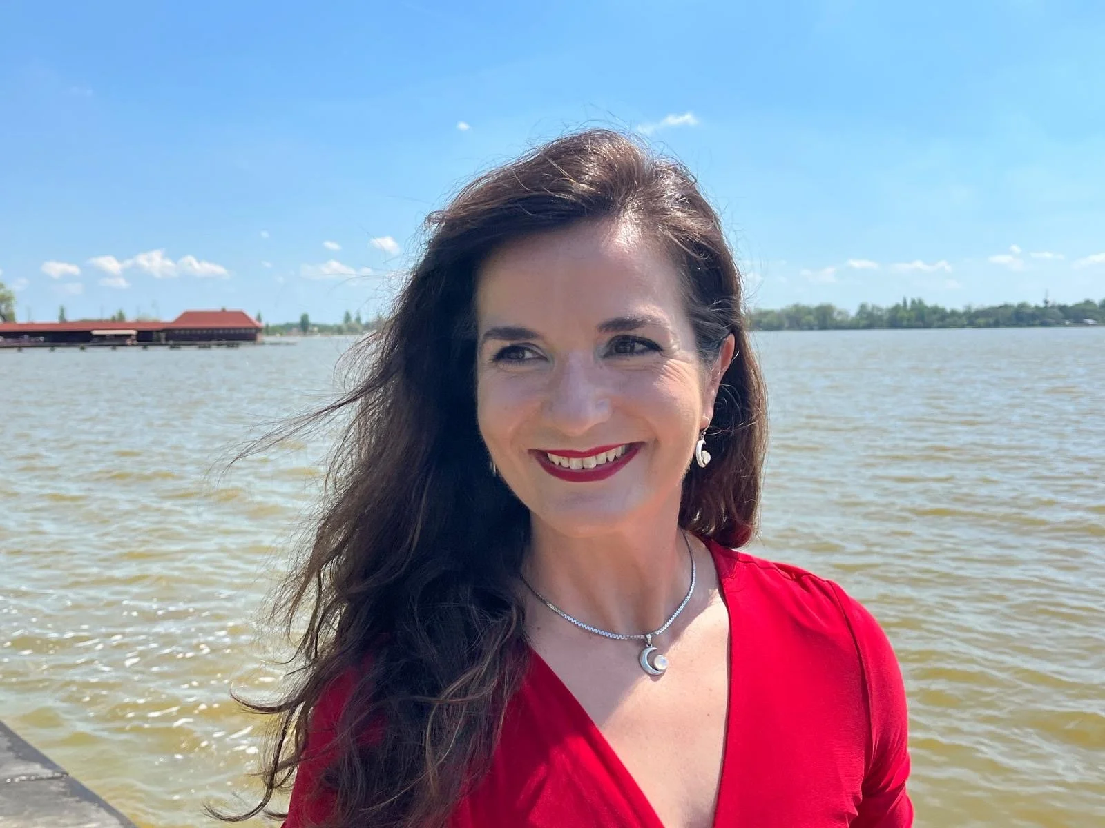 A woman with long dark hair smiling near a body of water under a sunny sky, wearing a red dress, earrings, and a necklace.