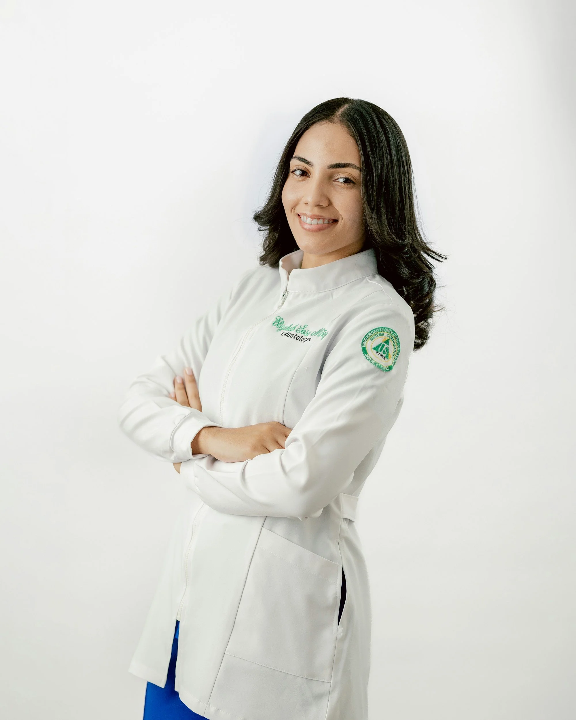 A young woman with dark hair in a white medical coat, standing with arms crossed and smiling, against a plain white background.