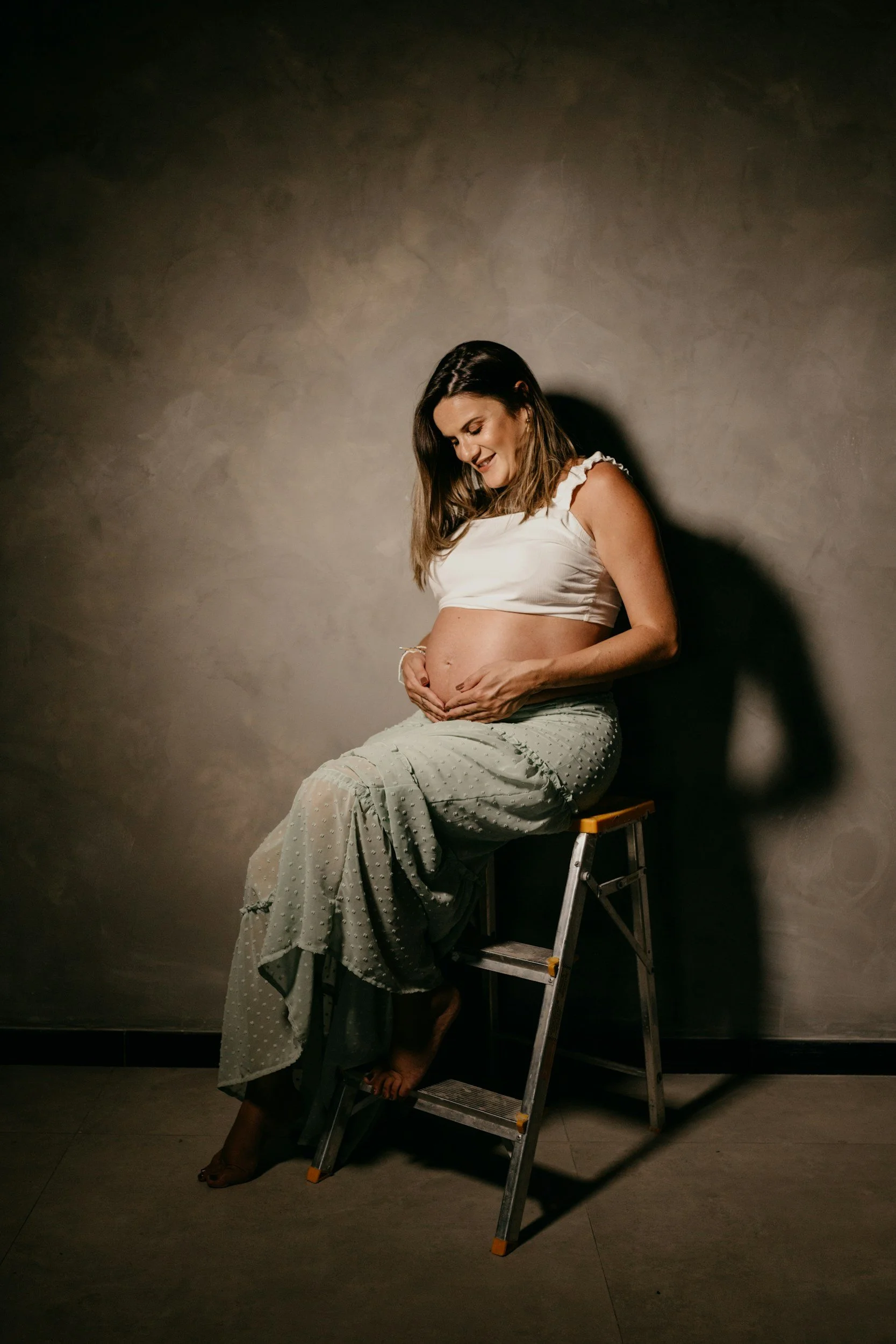 A pregnant woman sitting on a step stool against a plain wall, smiling and holding her belly.
