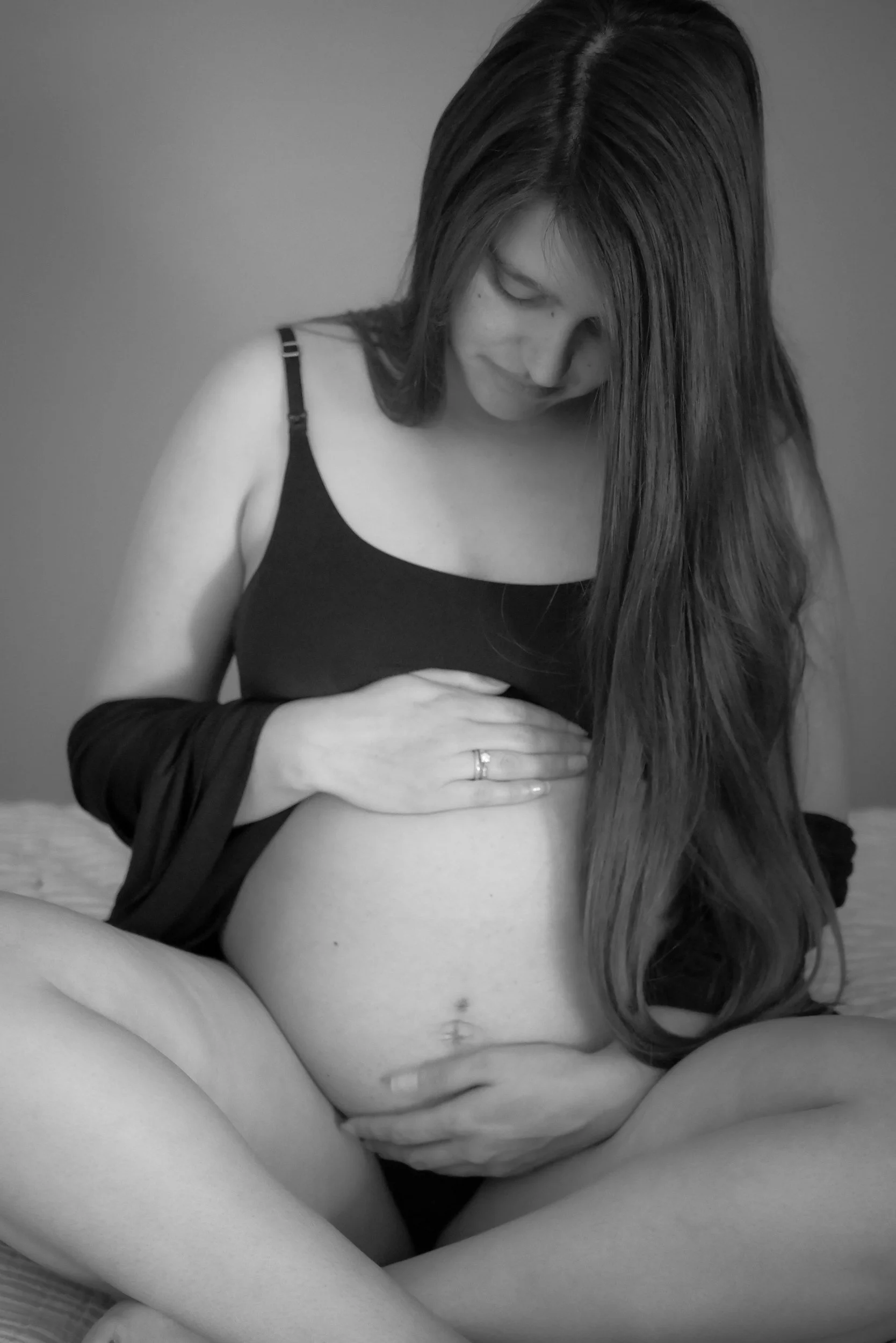 Black and white photo of a pregnant woman with long hair, sitting cross-legged, gently touching her belly, looking down.
