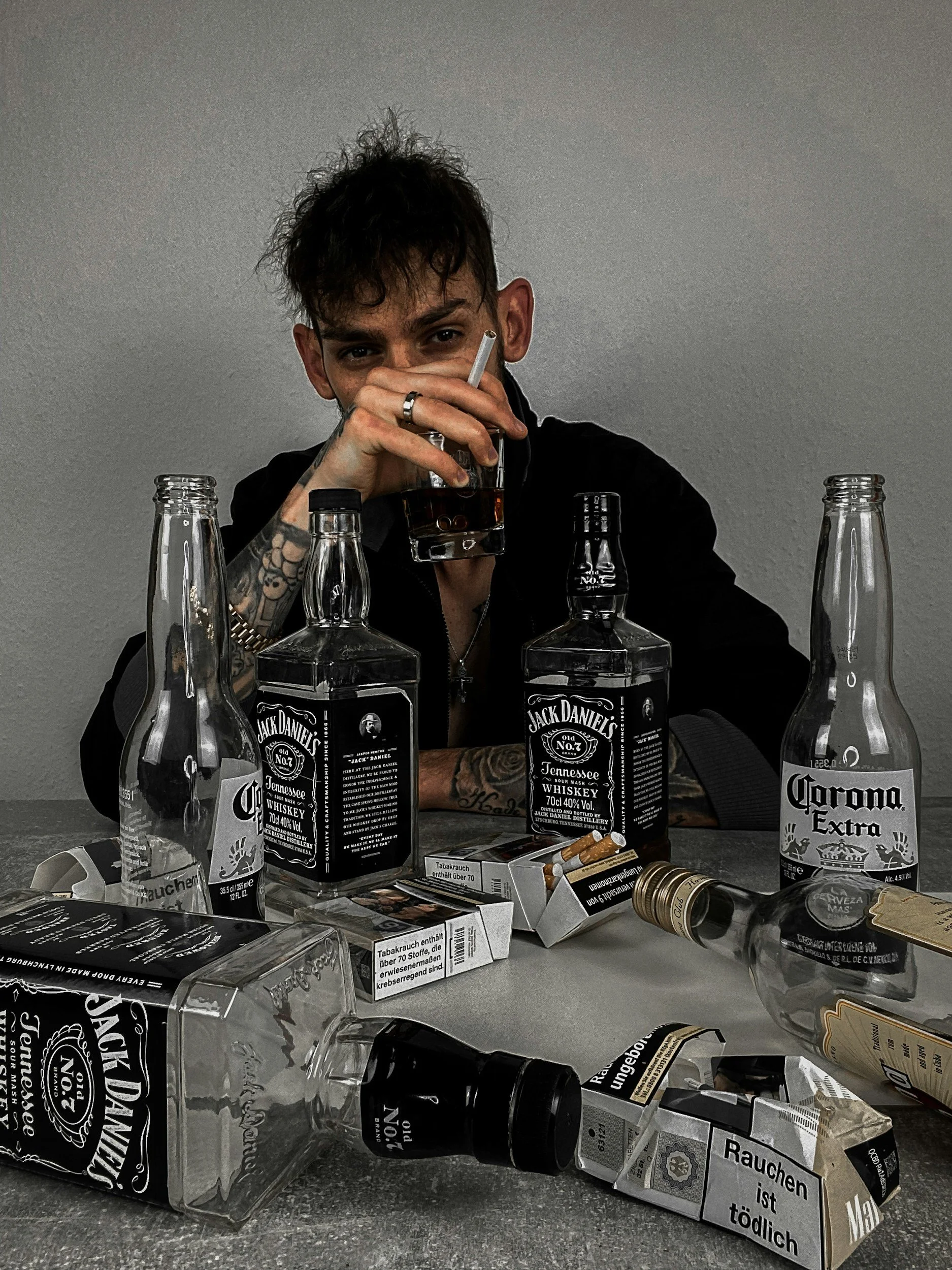 A young man with tattoos and dark hair sitting at a table cluttered with empty and full bottles of alcohol and cigarette packs, holding a glass of dark liquor with a cigarette in his hand, looking at the camera.