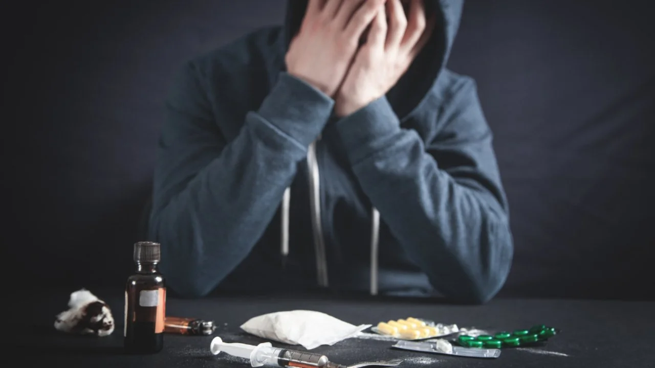 Person wearing a hoodie sitting at a table with drug paraphernalia including pills, a syringe, a bottle, and powder, suggesting drug addiction or overdose.