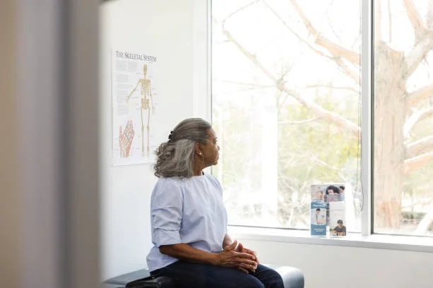 An elderly woman with gray hair sitting on a bench in a medical office, looking out a large window with trees outside, and a medical diagram on the wall.