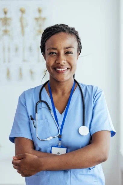 A female healthcare professional with a stethoscope around her neck smiling, wearing blue scrubs in a medical setting.