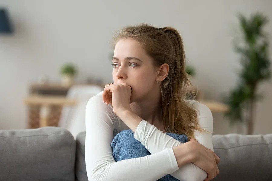 A young woman with brown hair in a ponytail sitting on a sofa, looking to the side with a contemplative expression, resting her chin on her hand.